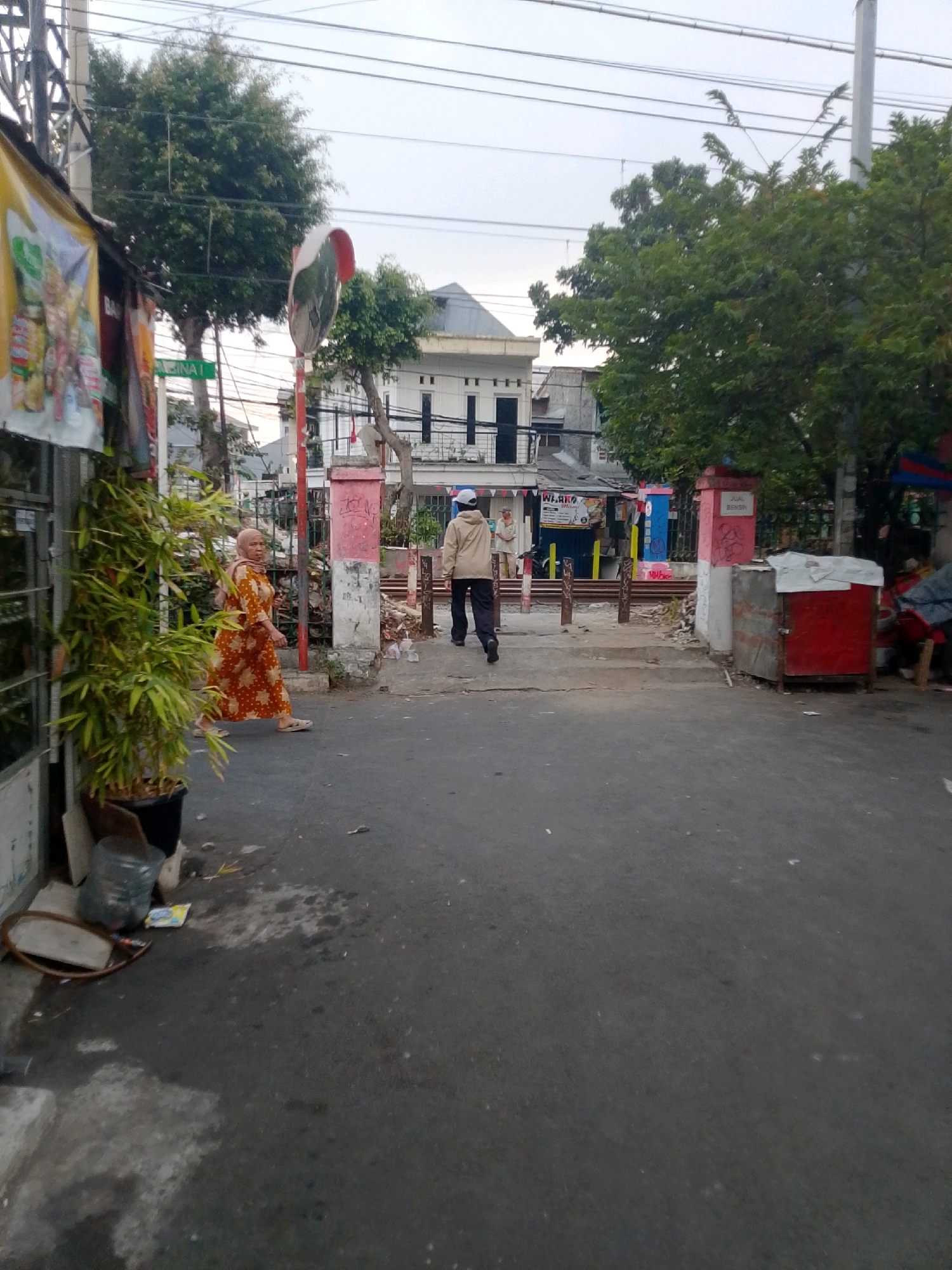 The image depicts a street scene in East Jakarta, Indonesia. In the foreground, a woman wearing an orange patterned dress and a headscarf is walking to the left. To her right, a man in a light-colored jacket and dark pants is walking away from the camera. Behind them, a street crosses railroad tracks, with a red and silver cart on the right side of the tracks. Buildings and trees line the street, and utility poles and wires are visible against a cloudy sky. A reflective traffic mirror is mounted on a pole to the left of the woman. The street is paved and appears to be in a moderately urban area.