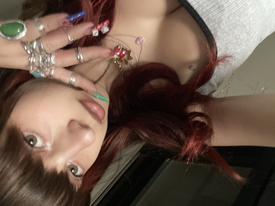 A woman with long, dark red hair and bangs poses, facing downward. Her hand, adorned with numerous rings and colorful, decorated nails, is raised near her face. She wears a light gray top. A necklace with various charms and beads is visible. Her fair skin, full lips, and light brown eyes are prominent. The image is taken from a close angle, focusing on her face and hand.