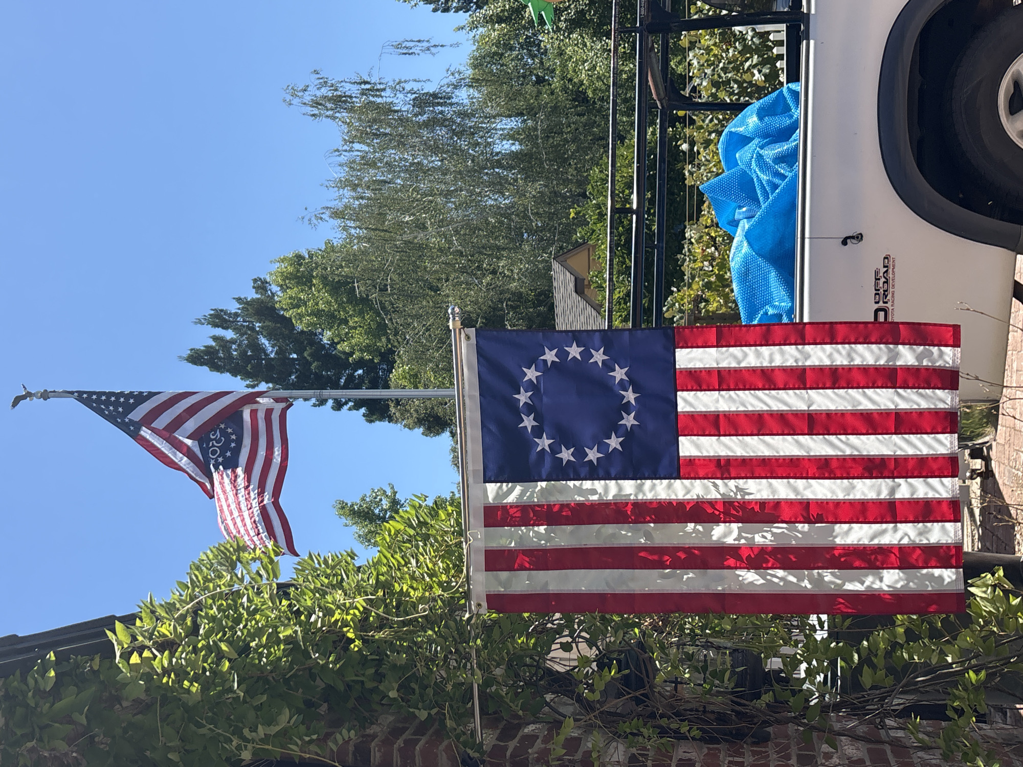 Two American flags are visible outdoors under a clear blue sky. One flag, likely a Betsy Ross variant with a circle of 13 stars, hangs from a pole attached to a brick wall on the left. The other flag, a more modern American flag with 50 stars, flies higher on a pole to the right, its stripes appearing somewhat distressed. Lush green foliage from a vine covers part of the brick wall. In the background, a white pickup truck with "OFF ROAD" and "TOYOTA RACING DEVELOPMENT" decals is parked. A bright blue, possibly pool cover, is folded in the truck bed. Trees with dense green canopies are scattered throughout the background, and a portion of a residential roof is visible. The overall scene suggests a sunny day in a residential or semi-rural setting. The location is identified as Citrus Heights, United States of America.