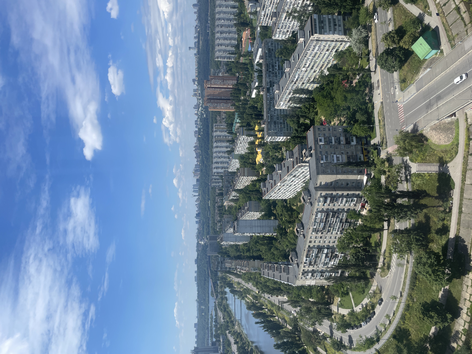 The image presents an aerial view of a dense urban landscape with a river running through it, characteristic of Kyiv, Ukraine. The city is characterized by numerous Soviet-era apartment blocks, many of which are monolithic and grey, interspersed with green spaces and trees. A wide river, likely the Dnipro, occupies the left side of the frame, with bridges crossing it in the distance. Roads with car traffic are visible throughout, with some curving through residential areas. The sky is blue with scattered white clouds, suggesting a fair-weather day. In the far distance, a more modern skyline with taller buildings can be seen. The overall impression is of a well-established city with a mix of older and newer architecture, nestled alongside a significant natural water feature.