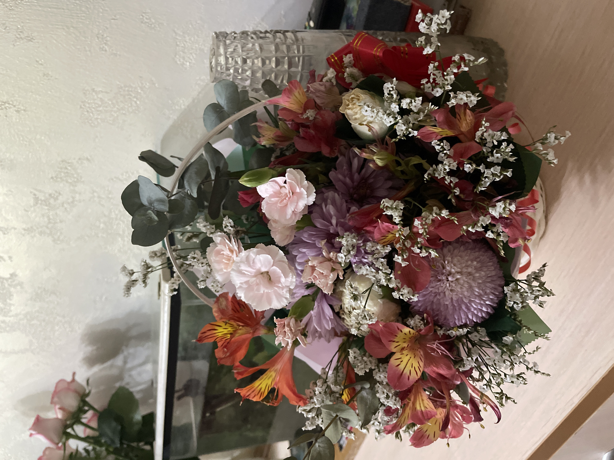A basket of flowers sits on a light wood surface. The basket contains a variety of colorful flowers, including orange and yellow alstroemeria, pink and white carnations, and purple chrysanthemums. There are also small white flowers and green eucalyptus leaves interspersed throughout the arrangement. A red ribbon with gold trim is tied around the basket. In the background, there is a white textured wall. To the left of the flowers, there is a partial view of pink roses in a vase, and a glass aquarium. To the right of the flowers, there is a tall, clear glass vase with a textured pattern. The lighting is dim, suggesting an indoor setting. The image does not provide any specific geographical markers for Almaty, Kazakhstan.