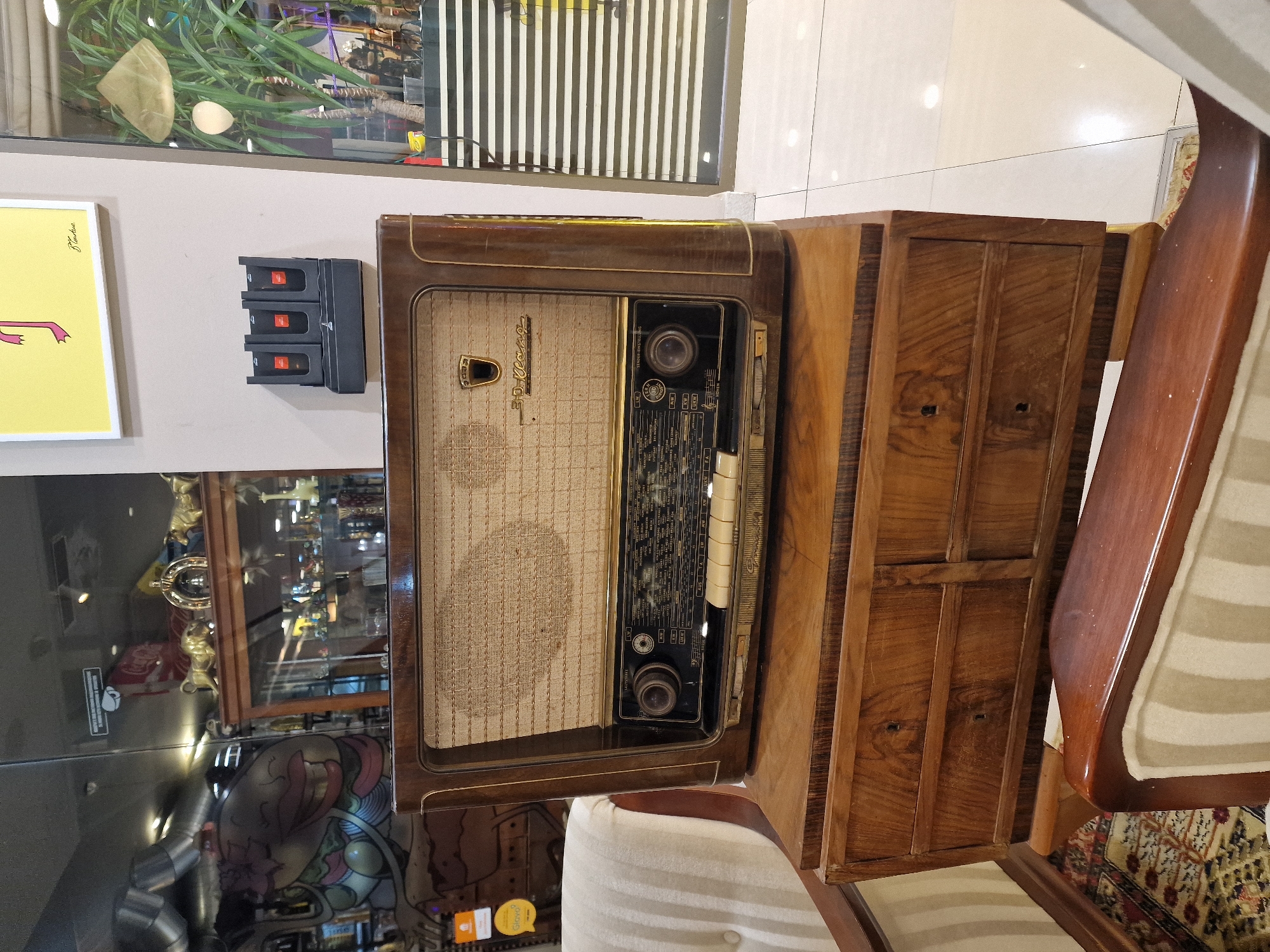 An antique wooden radio with a fabric speaker grill sits atop a wooden chest of drawers. The radio has a dial with station names and numbers, and several buttons. Above the radio, a black wall-mounted unit holds three items with red accents. The scene is indoors, with a tiled floor and a portion of a striped upholstered chair visible in the foreground. In the background, through a glass partition, various items are displayed, including a framed yellow artwork with a pink illustration, a large potted plant, and what appears to be a store interior with display cases and signage.