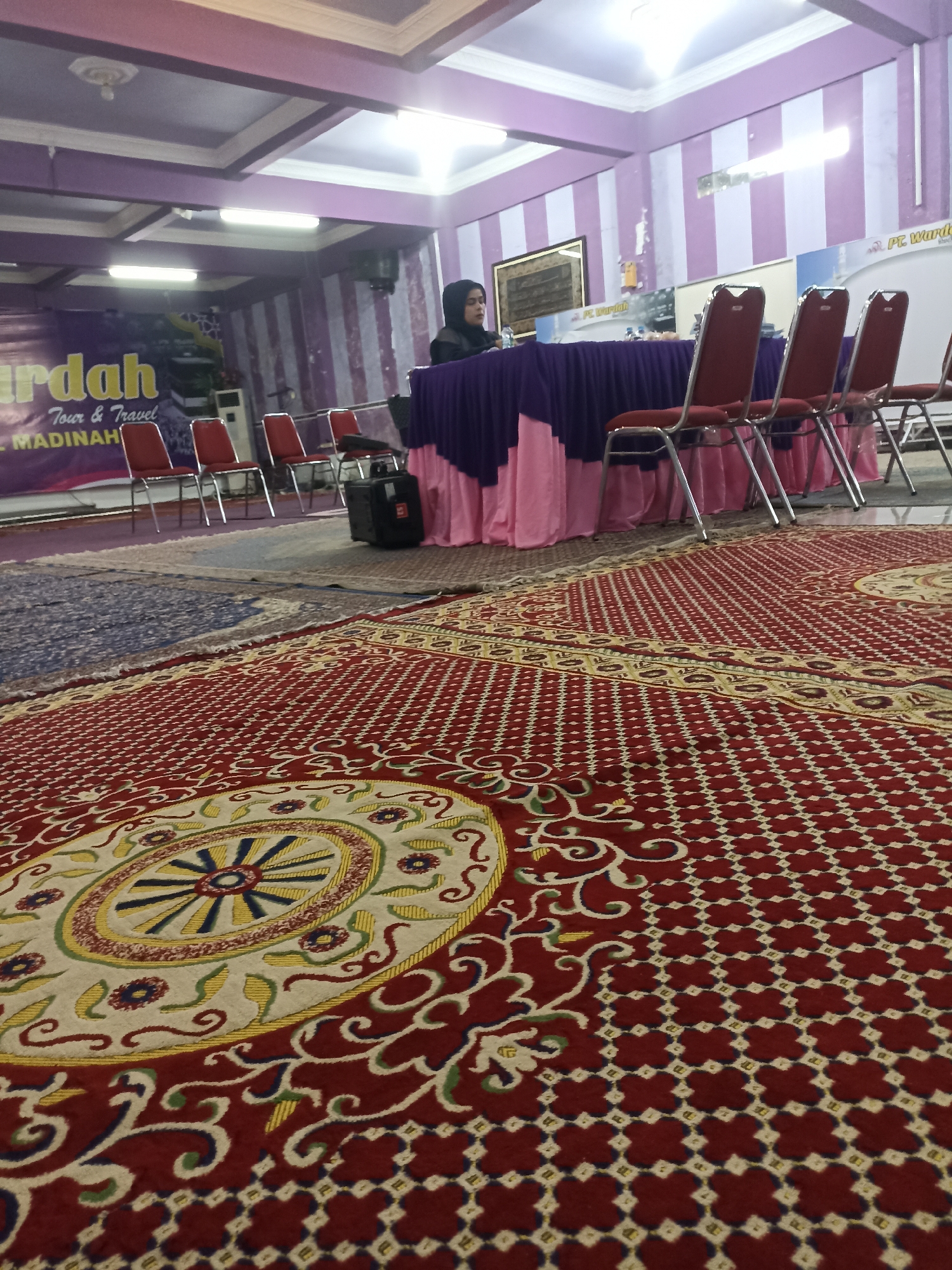 The image depicts an indoor scene, likely a conference or meeting room. A woman wearing a hijab is seated behind a table covered with a purple cloth and a pink skirted tablecloth. Several red chairs with metal frames are arranged in front of the table. In the background, a banner with the text "Murdah Tour & Travel" and "MADINAH" is visible. The walls are decorated with alternating purple and white stripes. The floor is covered with elaborate red and cream-colored patterned carpets. The ceiling has a suspended design with light fixtures. To the right of the scene, there is a sign that reads "PT. Wardah". The overall ambiance suggests a formal gathering or event. The specific location within Tompokersari, Indonesia, is not ascertainable from the visual information provided.