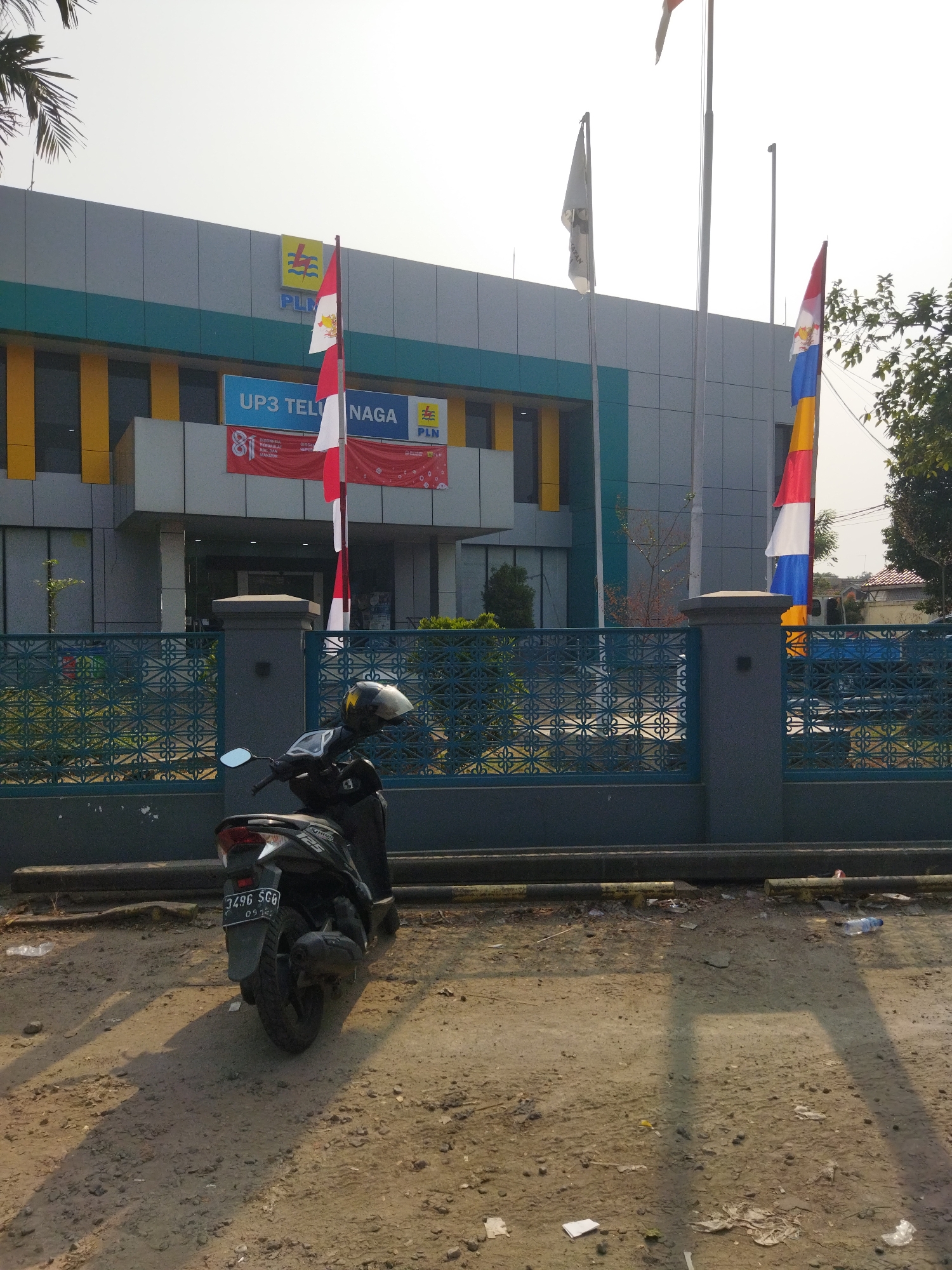 In the city of Pisangan Jaya, Indonesia, a dark-colored scooter is parked in front of a building that appears to be a PLN (Perusahaan Listrik Negara) office. The building has a modern facade with blue and grey panels, accented by yellow vertical stripes. Several flags are displayed outside, including Indonesian flags and a multi-colored flag with red, white, and blue sections. A banner above the entrance reads "UP3 TELUK NAGA". A blue metal fence with an intricate pattern separates the building from the foreground where the scooter is parked. The ground is unpaved and appears to be a mix of dirt and gravel, with scattered debris. The scene is illuminated by natural daylight, casting shadows on the ground.