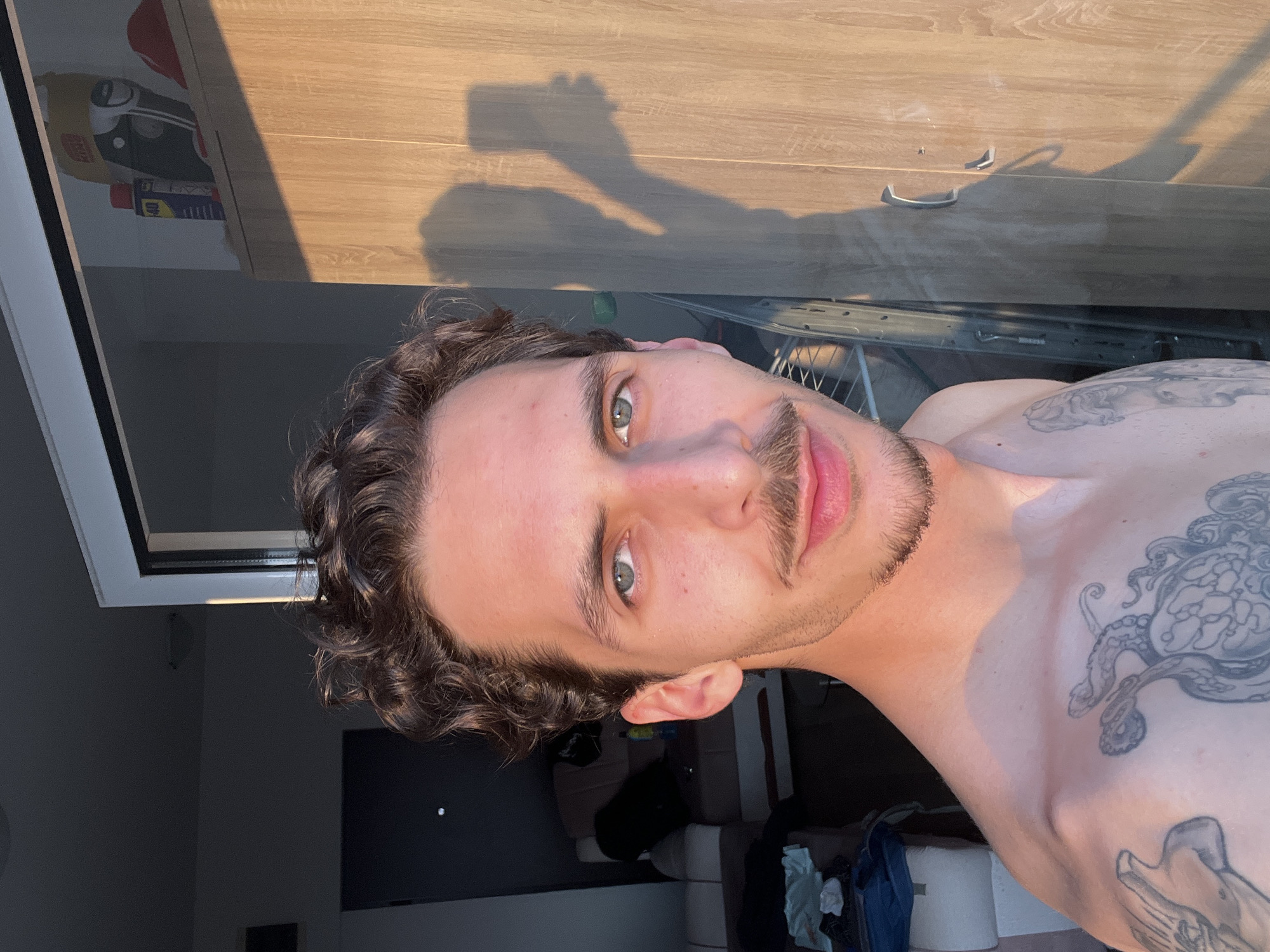 A man with curly dark hair and a mustache is taking a selfie. He is shirtless and has tattoos on his chest and shoulder. Behind him are partially visible objects: a wooden cabinet with metal handles, a window with a white frame, a dark room with a TV, and some clothes. The location is likely indoors, and the time of day appears to be sunny. There is no confirmation that the location is in the city of Bečići, Montenegro.