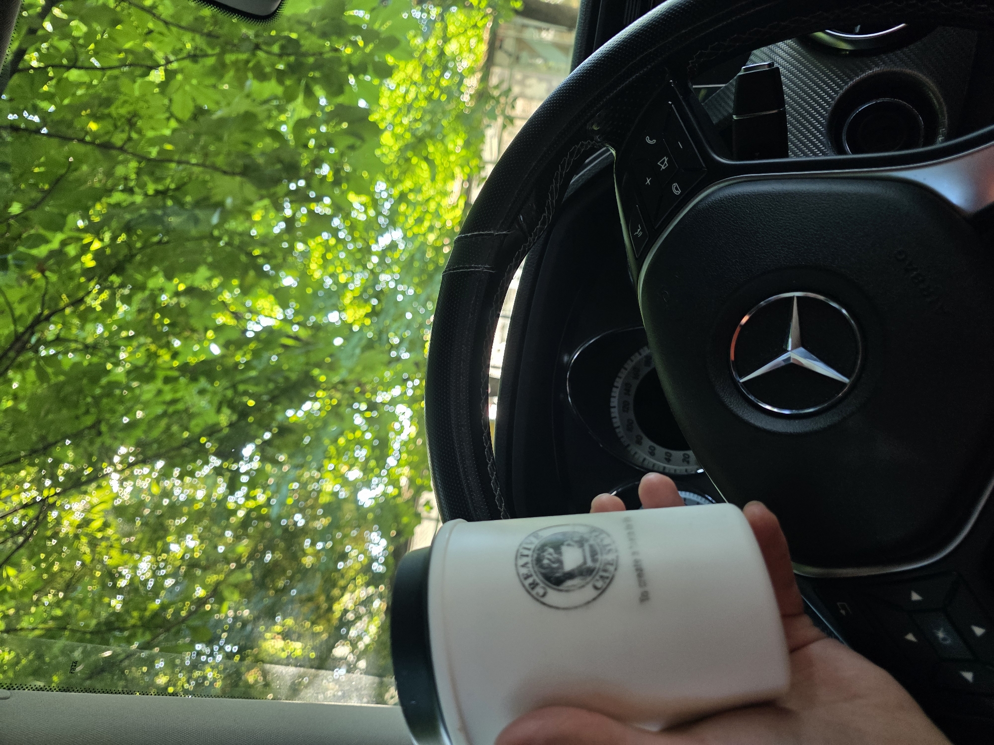 A person's hand holds a white paper coffee cup with a black lid, displaying a logo for "CREATIVE CAFE STYLE TO CREATE A BETTER LIFE" in front of a car's steering wheel. The steering wheel, which appears to be from a Mercedes-Benz, has various buttons and controls. Through the car's windshield, lush green leaves of a tree are visible, with sunlight dappling through them, creating a bokeh effect. The scene suggests a moment of pause or enjoyment while seated in a car, possibly parked in an outdoor location in Plovdiv, Bulgaria, surrounded by nature.