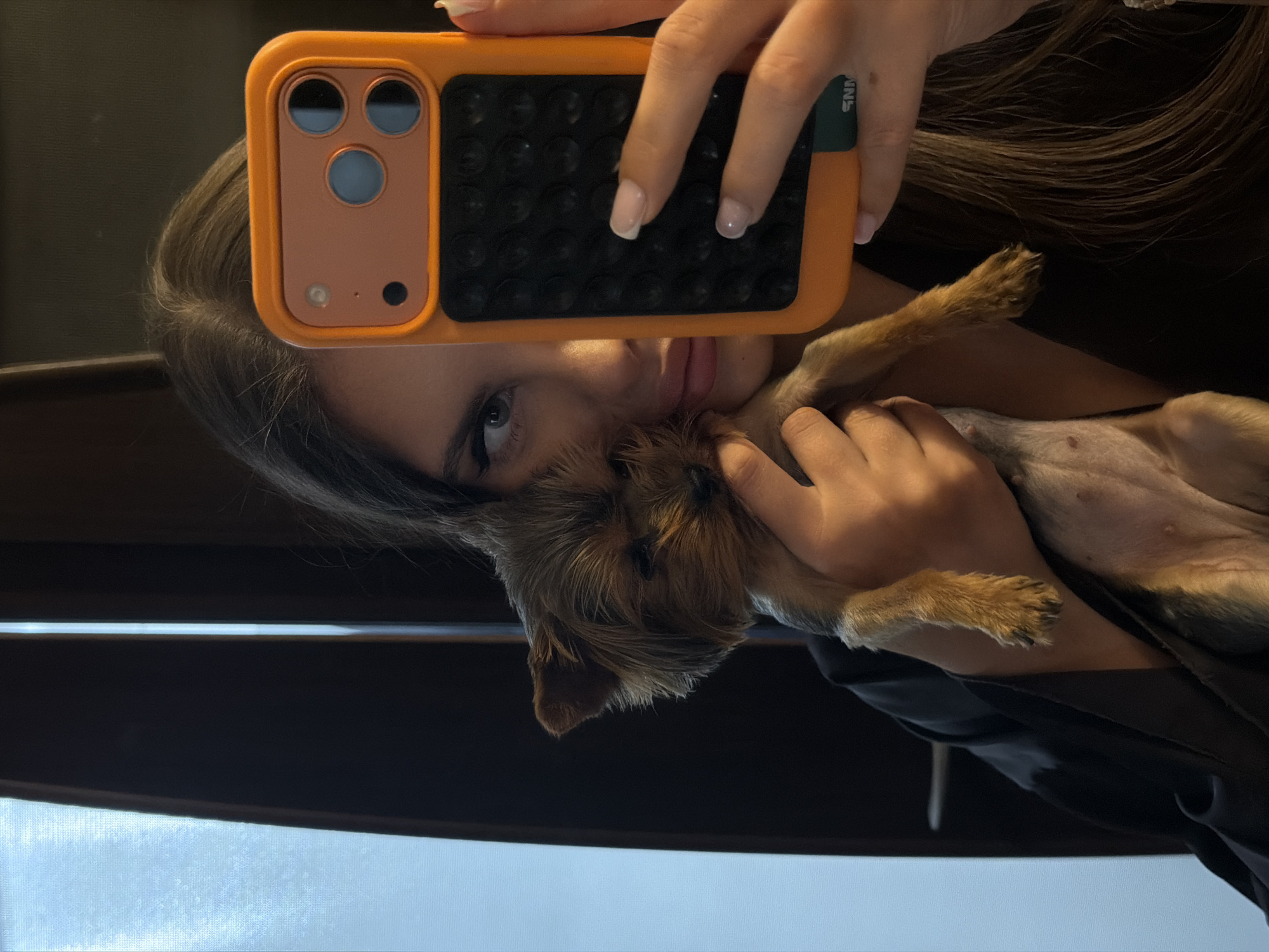 A selfie captures a woman holding a small brown Yorkshire Terrier-type dog. The woman's face is partially obscured by an orange and black phone case, but her eye is visible above. She has long brown hair. The dog's belly is visible. The location cannot be verified based on the image.