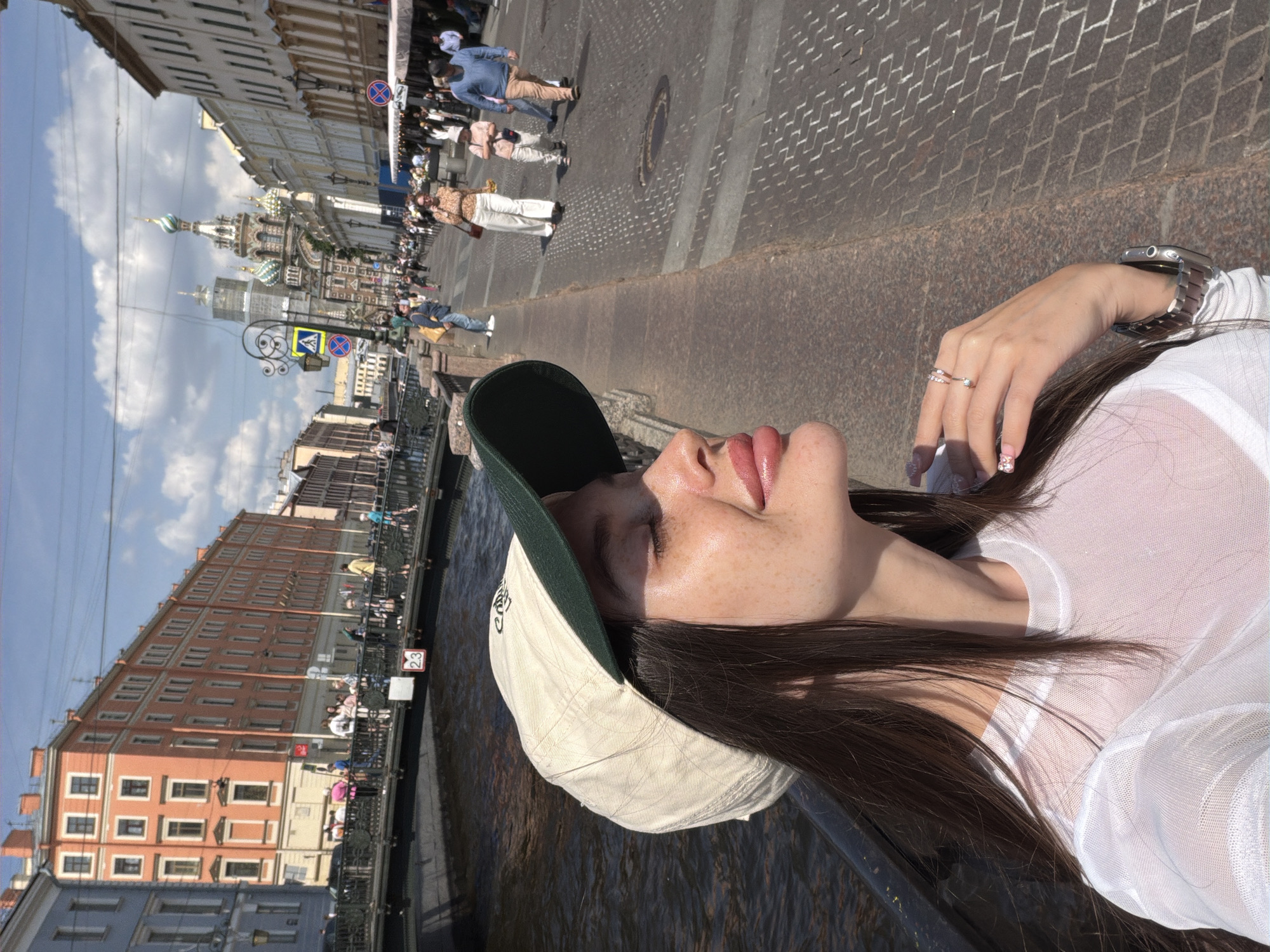 A young woman with long, dark hair and a light-colored baseball cap with a dark green brim is in the foreground of the image, looking to her right with her eyes closed and face tilted towards the sun. She is wearing a sheer white long-sleeved shirt. Her right hand, adorned with rings and decorated fingernails, rests near her chin. A silver watch is visible on her left wrist. She is standing on a stone embankment next to a canal.
In the background, a street scene unfolds along the canal. A pedestrian bridge spans the canal, with several people walking across it. Buildings line the opposite side of the canal, including a prominent orange building with numerous windows and a light blue building.
Further down the street, on the right side of the image, a paved walkway is filled with pedestrians. Shops with awnings are set up along this walkway. In the distance, the iconic onion domes of the Church of the Savior on Spilled Blood are visible, its colorful facade rising against a blue sky with scattered clouds. Street lamps and traffic signs are also present along the street. The architecture suggests a historic European city, consistent with Saint Petersburg, Russia.