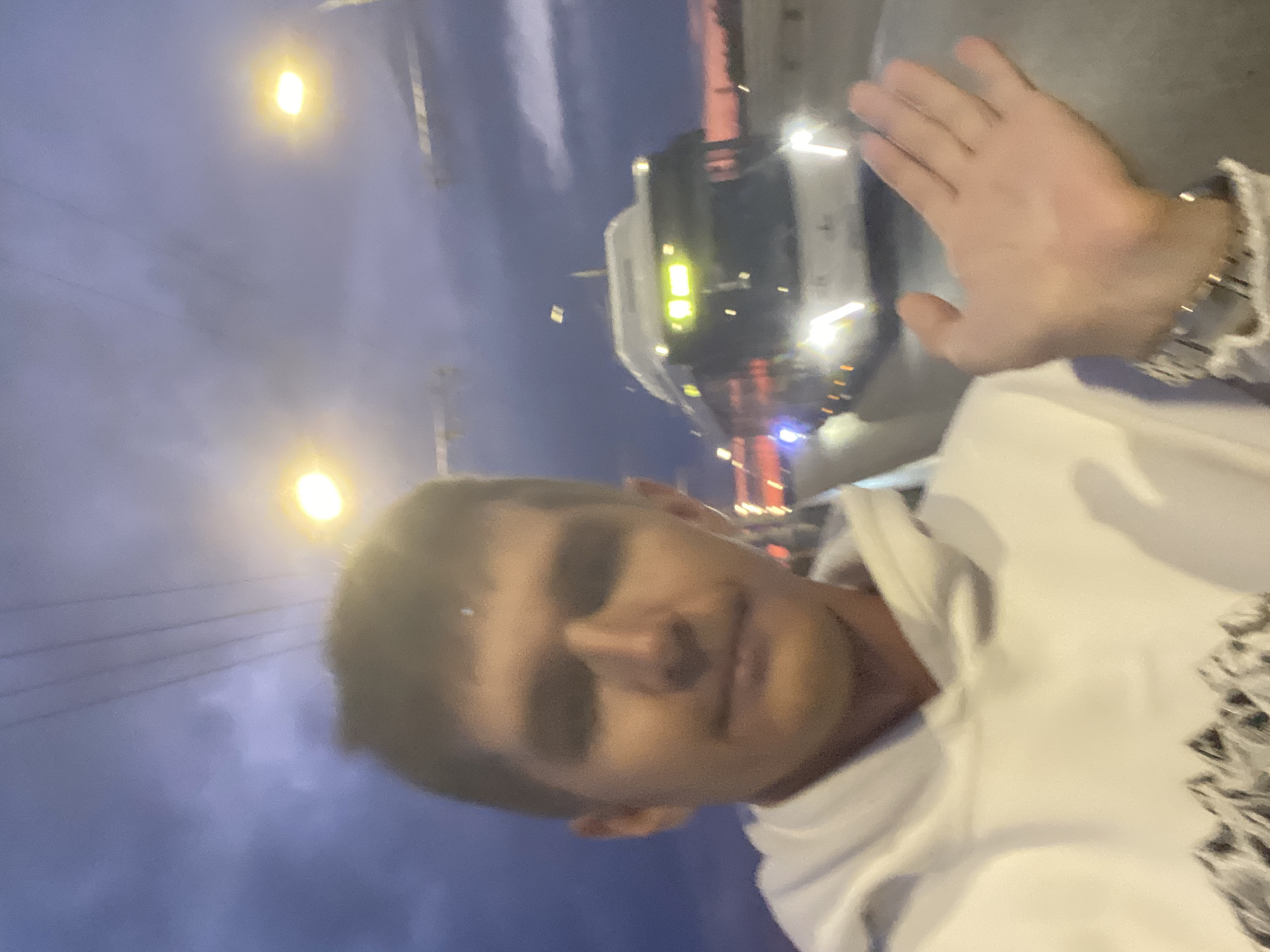 The image depicts a young man in a white hoodie taking a selfie outdoors at dusk. He is standing on a road, with a bus in the background. The sky is cloudy with hints of sunset colors. Streetlights are visible, casting a yellow glow. The man's left hand is raised, and he is wearing a watch on his left wrist. The bus appears to be a modern public transport vehicle, possibly a trolleybus, with illuminated headlights and destination display. The overall setting suggests a street scene in an urban or suburban area. The specific location in Novokuybyshevsk, Russia, is not discernible from the visual information provided in the image.