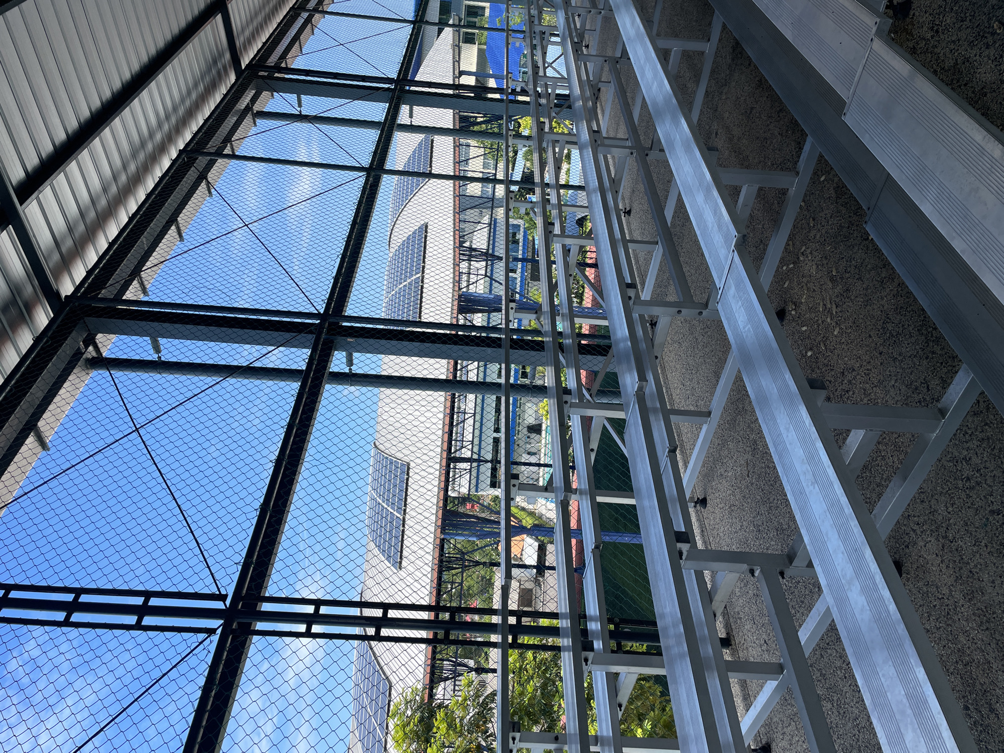The image shows rows of empty silver bleachers under a covered walkway. A chain-link fence runs along the front of the walkway, providing a view of a building with solar panels on its roof. Trees and other buildings are visible in the background. The ground under the bleachers is paved with gravel. The sky is blue with some scattered clouds.