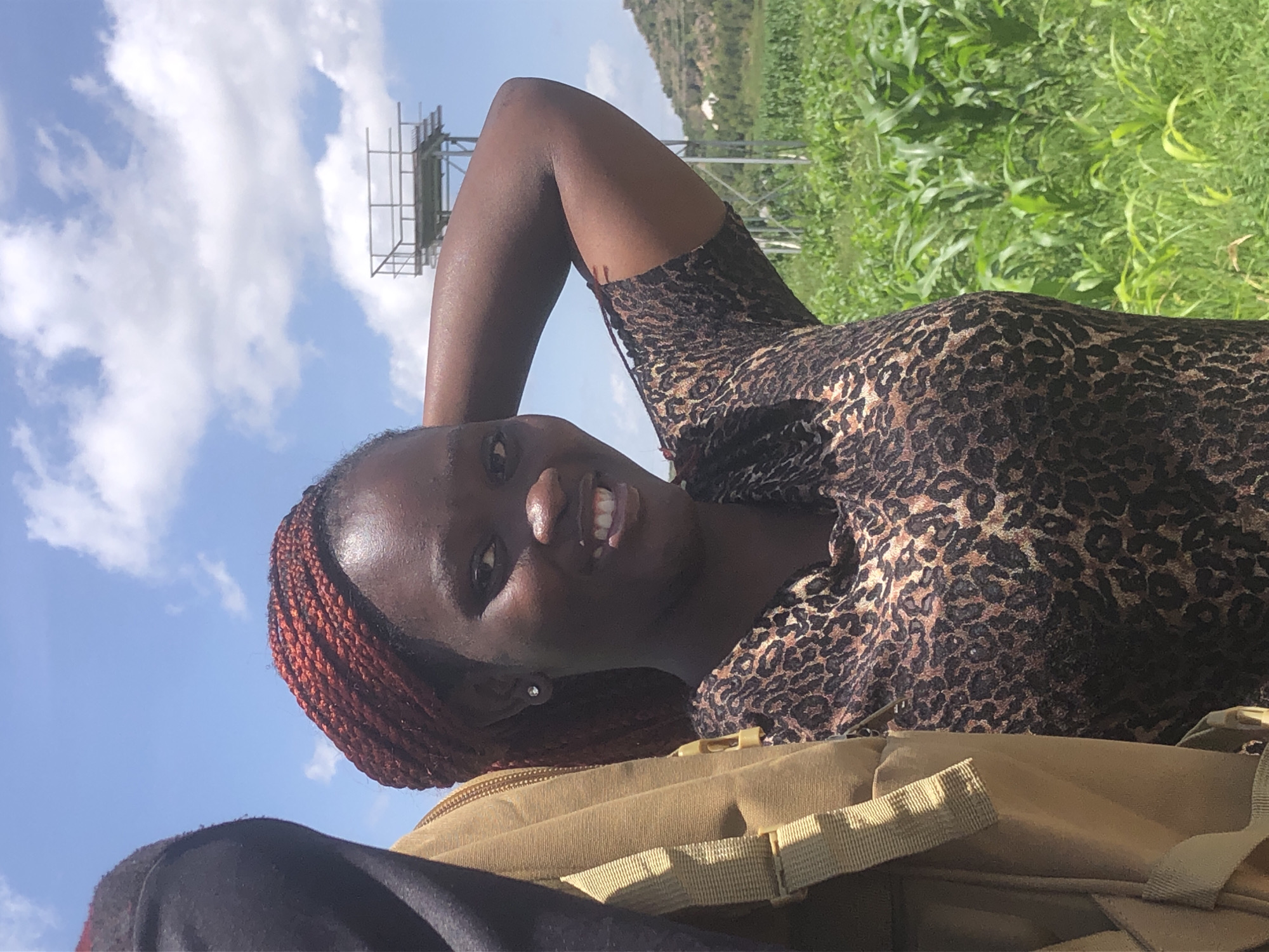 A medium shot captures a young Black woman with braided auburn hair and a leopard-print shirt smiling and posing outdoors. She is partially obscured by a tan backpack on the left side of the frame. Her right arm is raised and bent behind her head. In the background, a field of green corn stalks stretches towards a distant hill. A metal structure, possibly a water tower, is visible against a blue sky with white clouds. The scene is set in Gwafan, Nigeria.