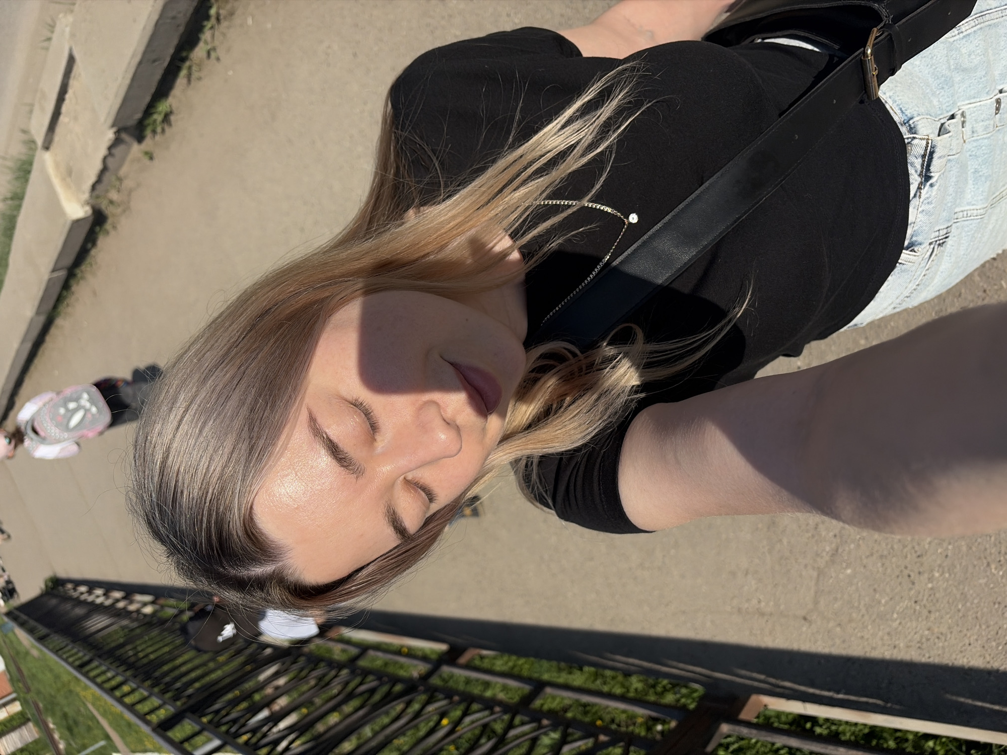 The image shows a woman in a black top and light blue jeans with a black shoulder bag, standing in Chaikovsky, Russia. She appears to be taking a selfie, with her eyes closed and face turned towards the camera. The background includes a sidewalk, a metal railing, and a patch of grass. Another person is visible in the background, walking with a child in a stroller.