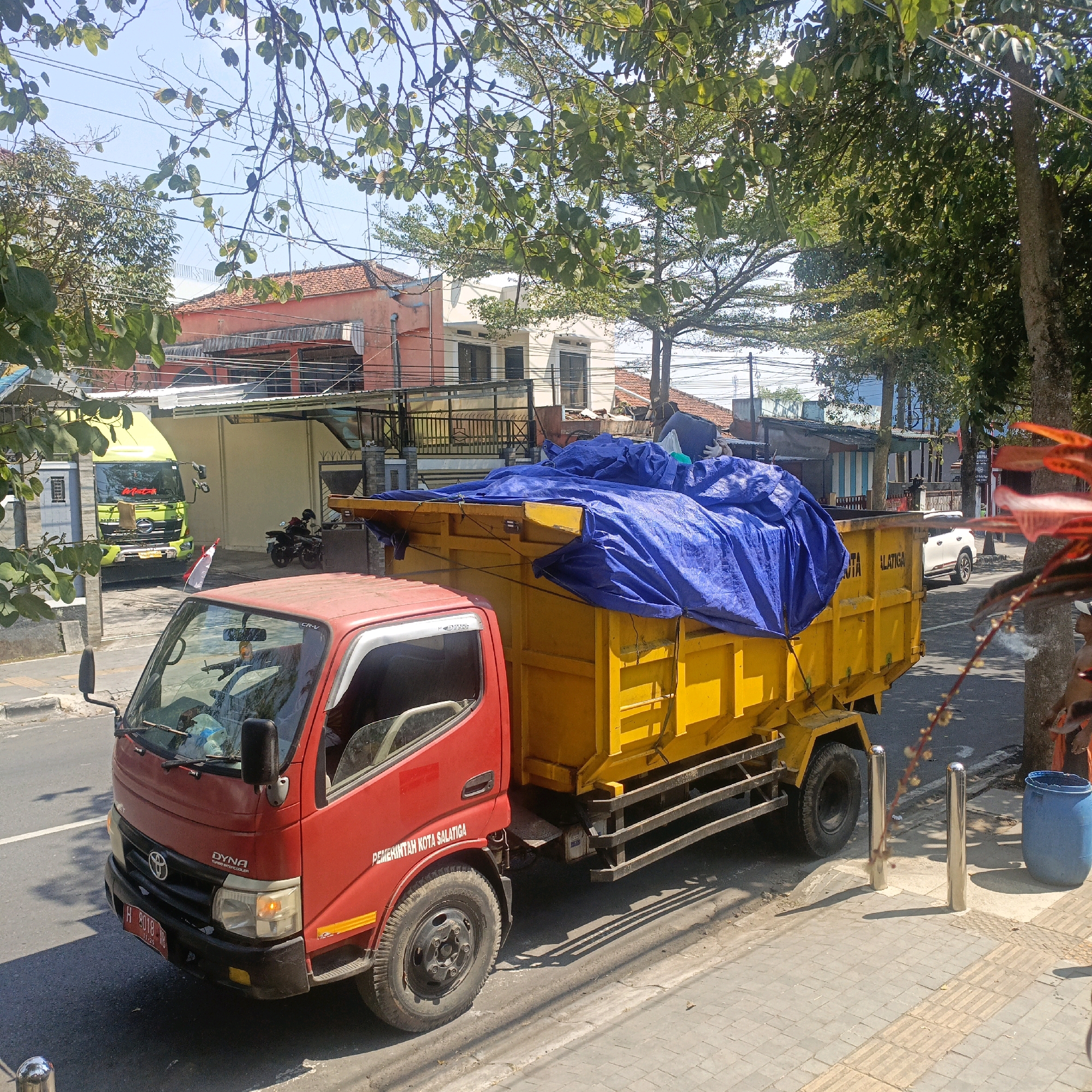A red pickup truck with a yellow dump truck trailer is parked on a paved street in Salatiga, Indonesia. The dump truck is covered with a blue tarp. A green truck is parked to the left of the dump truck. A motorcycle is parked in the background. Buildings are visible in the background. There are trees and branches in the foreground. The sky is blue.