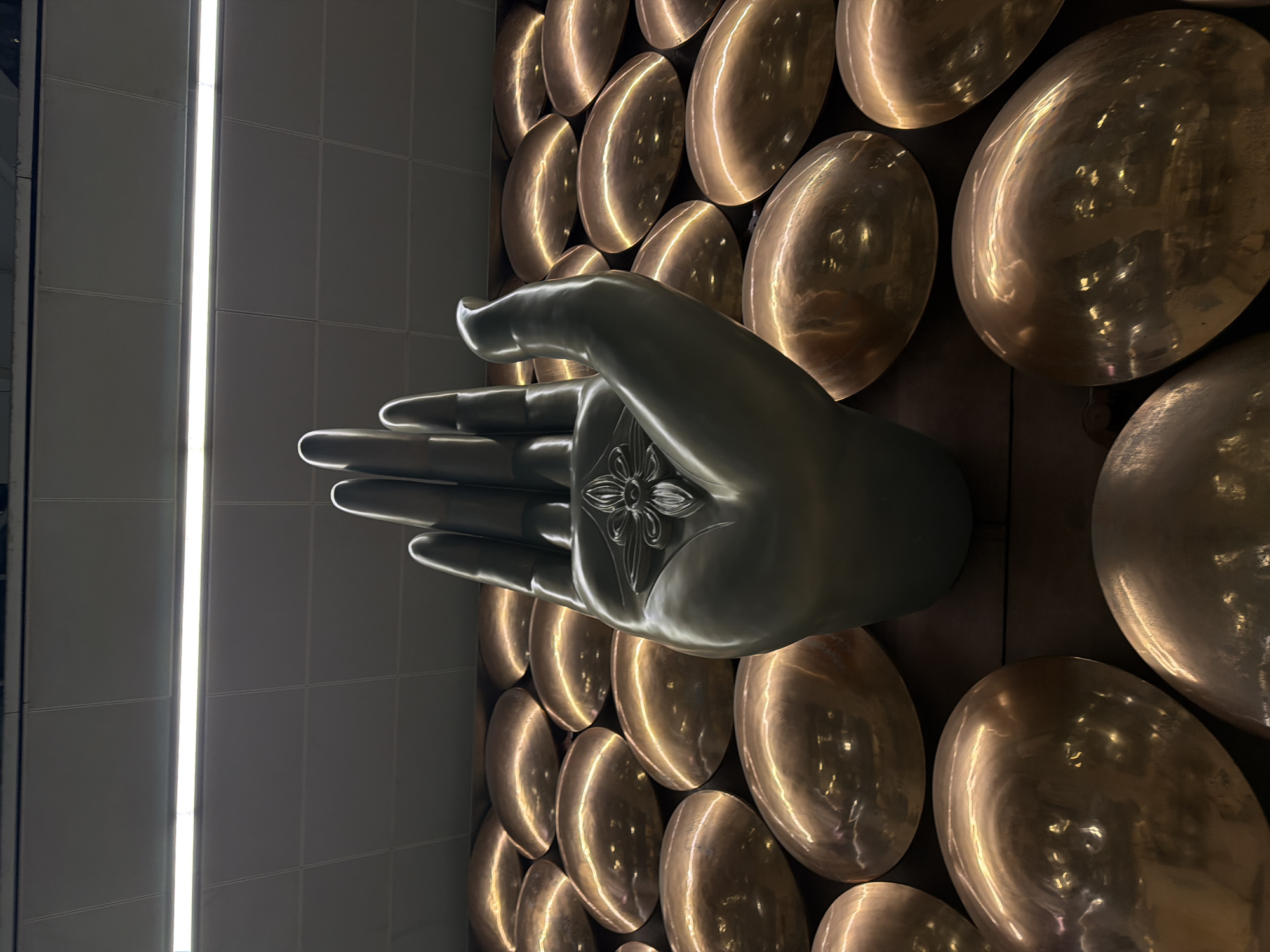 An imposing, metallic sculpture of an open hand, with fingers extended upwards, stands prominently in the foreground. The hand sculpture is dark gray, with a floral design embossed on the palm. Surrounding the hand, the wall is adorned with a mosaic of numerous large, oval, copper-colored objects, arranged in a tessellated pattern. These objects are highly reflective, catching and mirroring the ambient light. The ceiling above is a grid of square panels, with a long fluorescent light fixture running horizontally across the center, illuminating the scene. This artwork is located in the departure hall of Indira Gandhi International Airport (DEL) in Delhi, India.