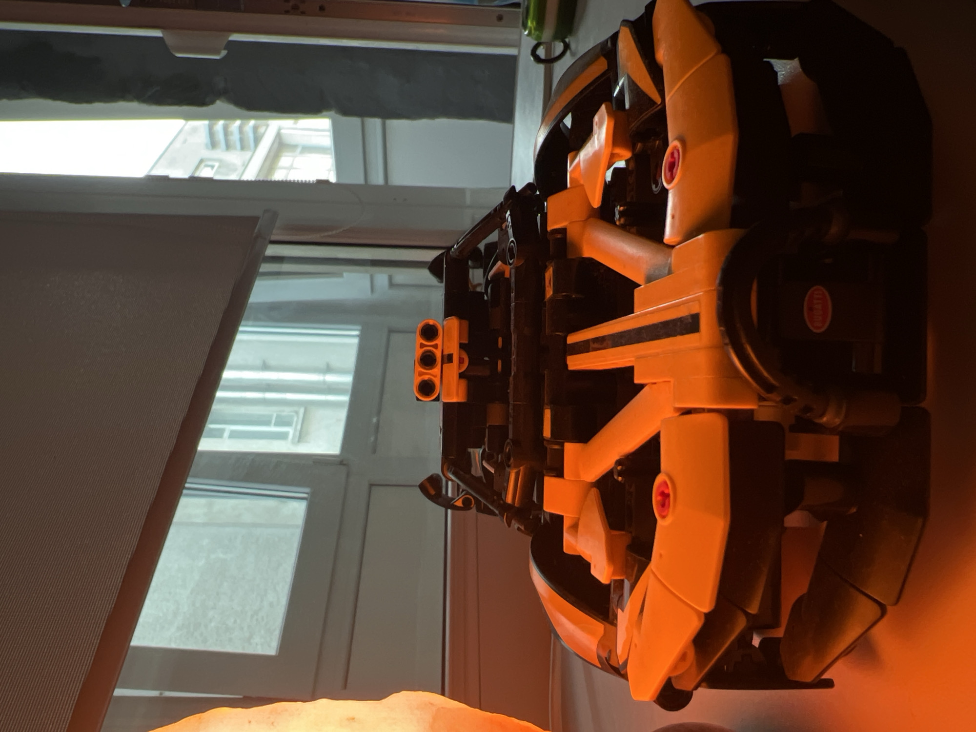 The image features a detailed, complex model, likely constructed from building blocks such as Lego Technic, rendered in black and yellow/orange hues. The model appears to be a sophisticated vehicle or machine, showcasing various mechanical elements including panels, beams, and a component with three circular openings, possibly representing cannons or exhaust pipes. A notable detail is a red circular sticker on a black part of the model, which clearly displays the word "BUGATTI."
The setting is indoors, with the model positioned on a surface. A warm, amber glow emanates from a light source, possibly a salt lamp, which is partially visible in the bottom foreground, casting an orange hue over parts of the model and its immediate surroundings. Behind the model, a white-framed window offers a view to the outside. Through the window, the light-colored facade of another building with multiple windows is discernible, suggesting an urban environment. A light-colored roller blind or curtain is partially lowered across the window. No individuals are present in the image, and no specific event or activity is taking place; the scene is a static display of the model.