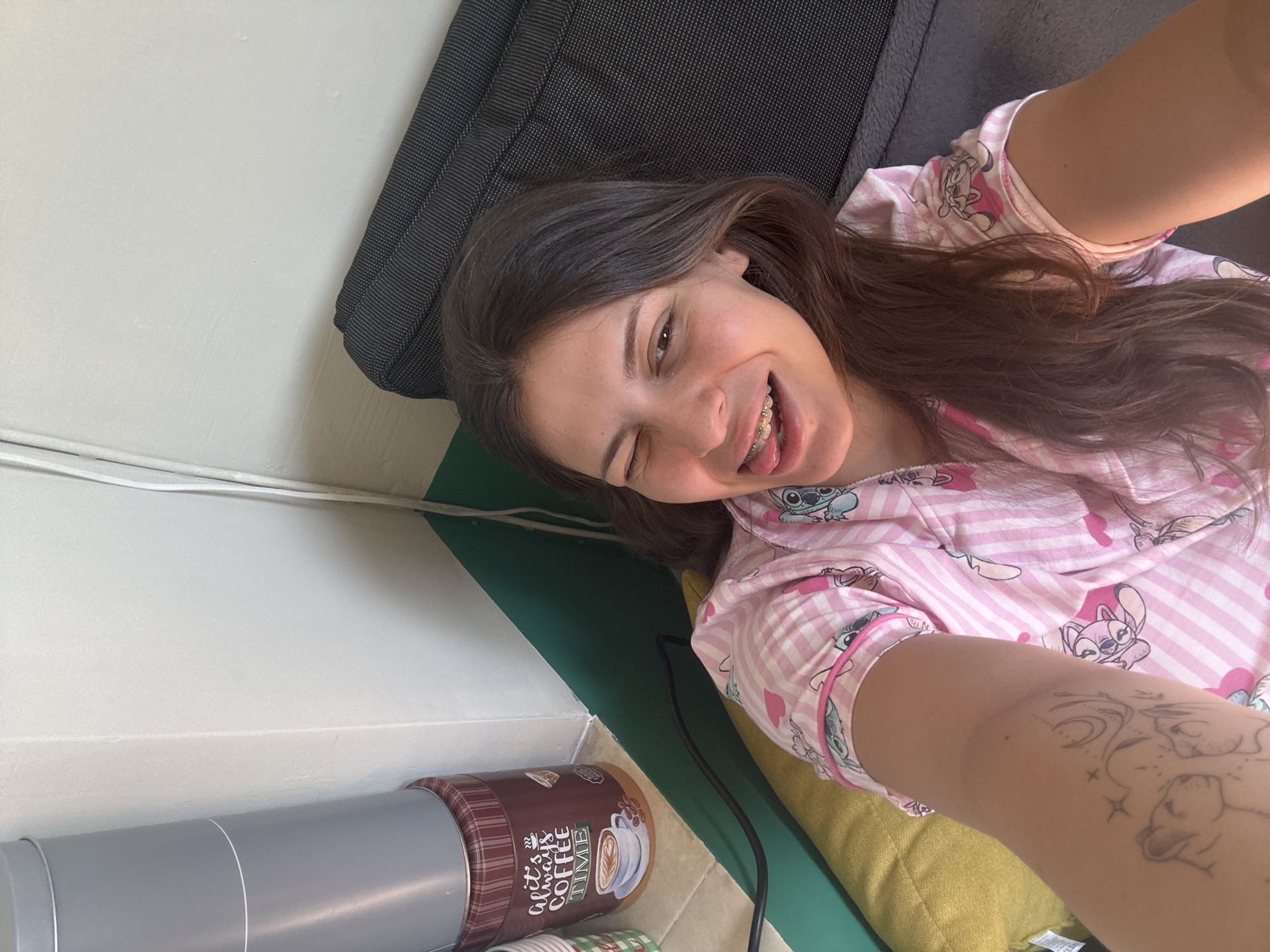 The image is a selfie taken from a high angle, showing a young woman with long, dark brown hair lying on a bed. She is wearing pink and white striped pajamas with a Stitch character print. She has braces on her teeth and is sticking out her tongue while winking. Her left arm, visible in the foreground, has a tattoo of two lion cubs looking up at celestial elements.
The woman is positioned on a yellow surface, possibly a mattress or cushion, next to a dark grey upholstered headboard. To the left of the frame, there is a tall, cylindrical grey object on top of a red and white checkered cylindrical container, which appears to be a coffee can. The background consists of a white wall with a green stripe along the bottom. The overall environment suggests a domestic setting, likely a bedroom.
Without further information or visual cues, it is impossible to determine the specific location within the city of Plovdiv, Bulgaria.