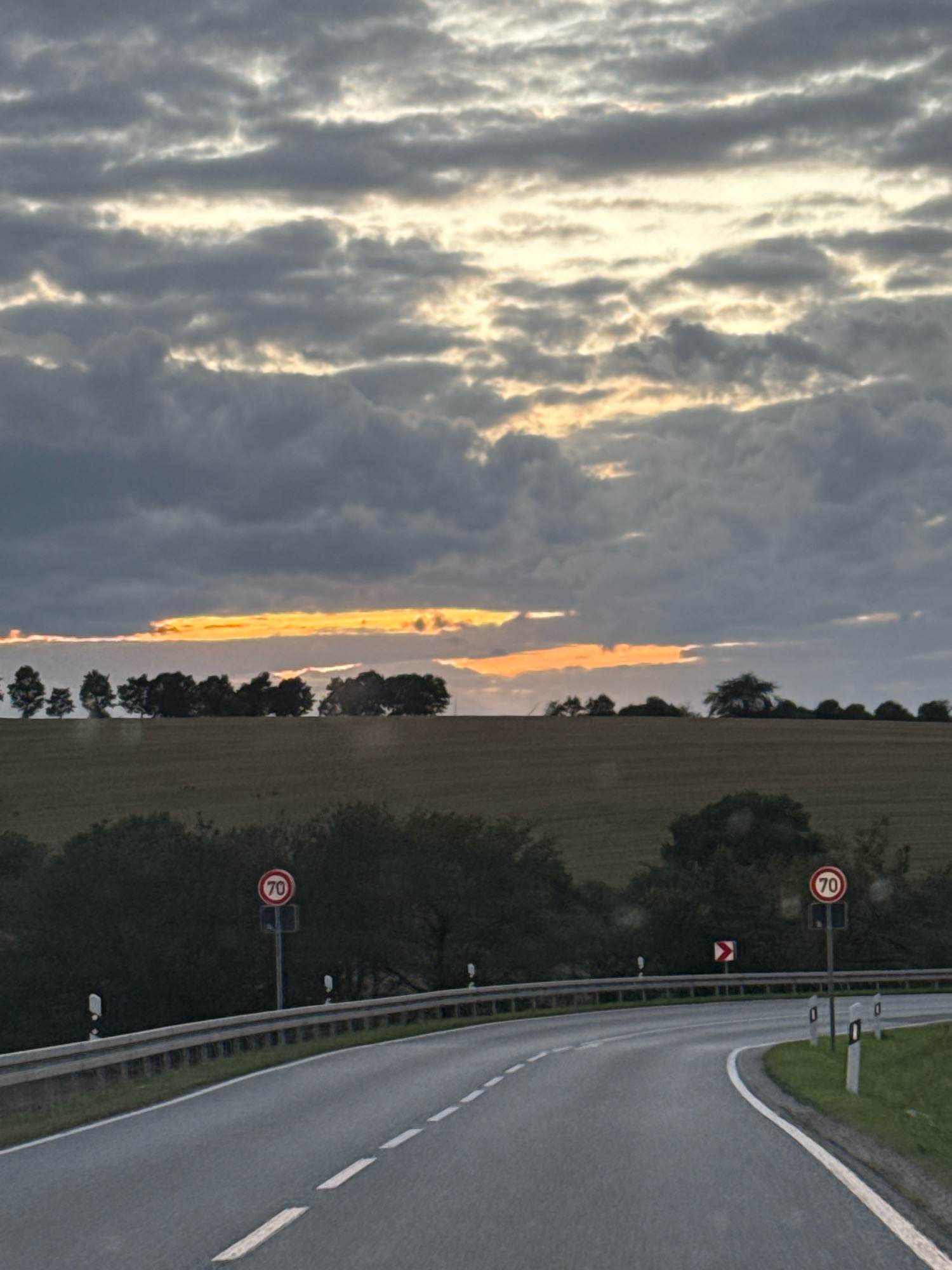 The image depicts a curved road in Nordhausen, Germany, with a cloudy sky and a hint of sunset. On the left side of the road, a guardrail runs along the edge, with several white reflective markers. A speed limit sign indicating "70" is visible on a pole, accompanied by another traffic sign showing a curved arrow. The road is asphalt with white dashed lines marking the lanes. On the right side of the road, there is a grassy embankment. In the background, a line of trees forms a dark silhouette against a backdrop of a brown field. The sky above is filled with dark, textured clouds, with a bright orange band of light peeking through them, suggesting either sunrise or sunset. There are no people visible in the image.