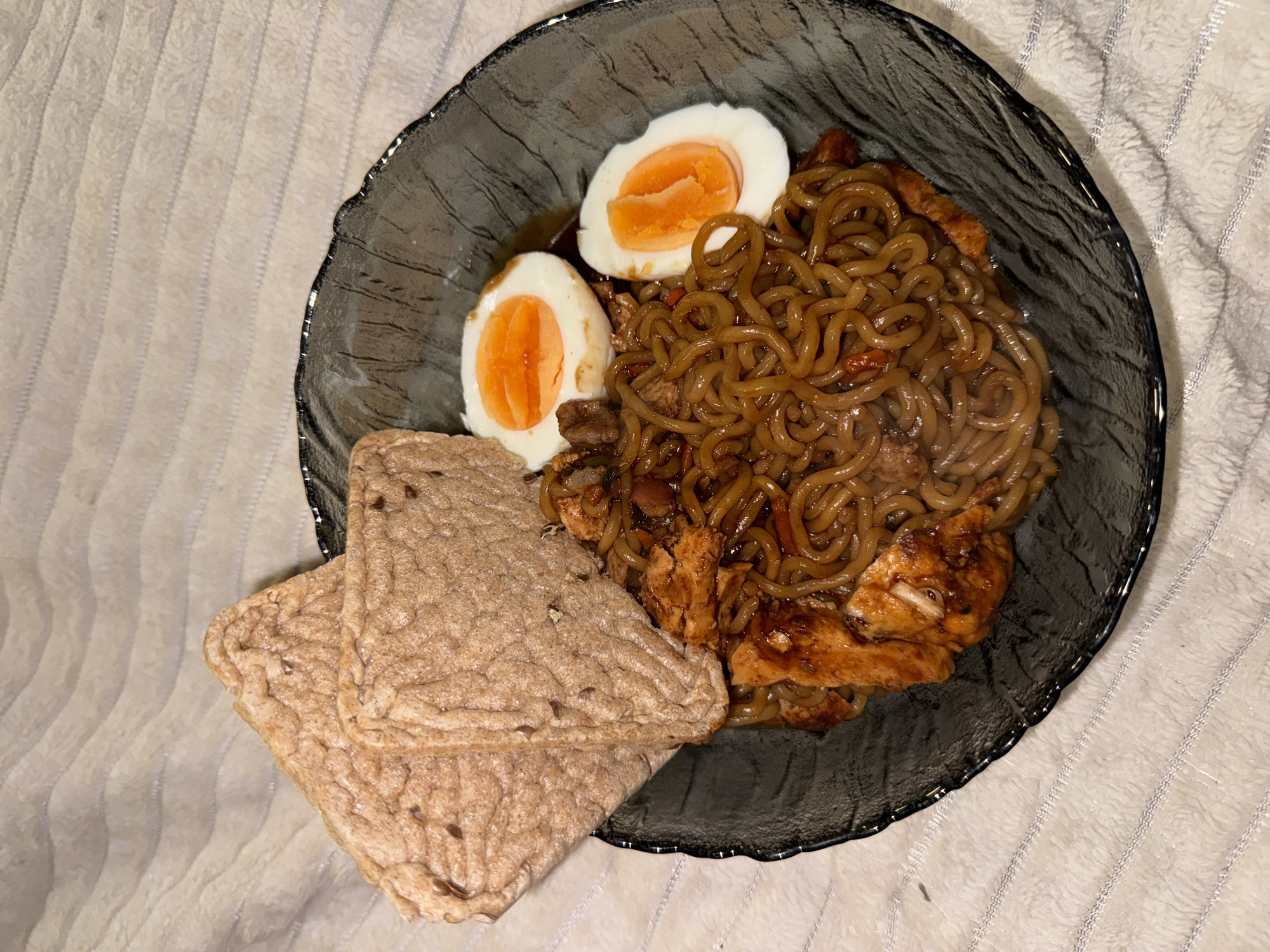 The image shows a black bowl filled with noodles and chicken, served with a sliced egg and two flatbreads. The bowl sits on a white surface with a textured pattern. The scene takes place in an unknown location.