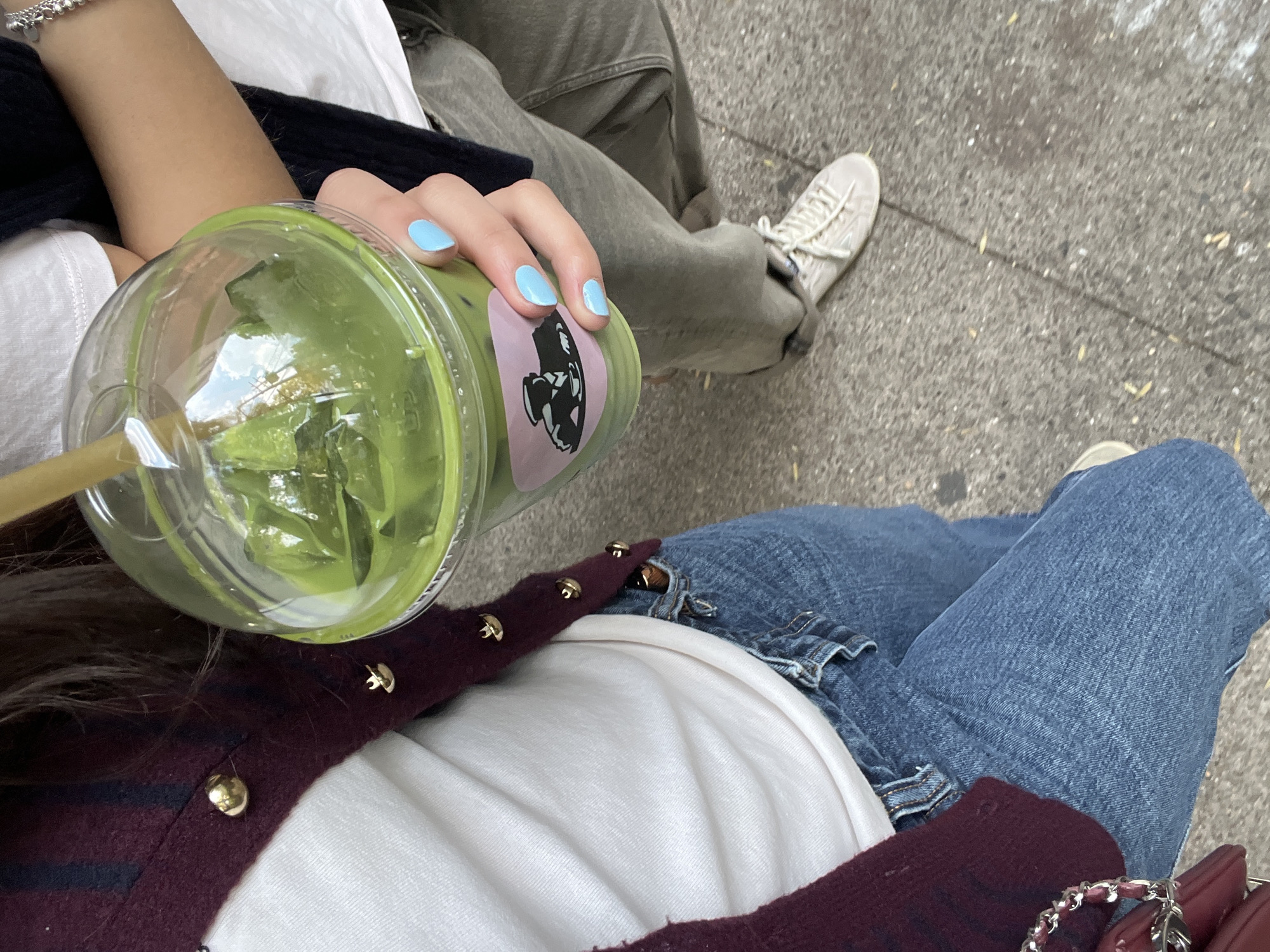 The image shows two people sitting outside in the city of Unknown, Unknown, possibly on a sidewalk. The person in the foreground is wearing a white t-shirt with a maroon cardigan that has gold buttons. They are wearing blue jeans. Their manicured hand, with pale blue nail polish, holds a large, clear plastic cup filled with a green beverage, likely matcha, and a straw. The cup has a sticker of a graphic image. The other person is wearing grey pants and off-white sneakers. The ground appears to be concrete.