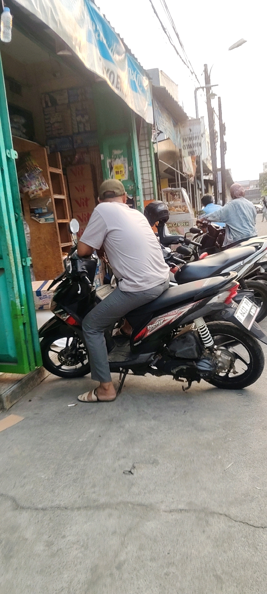 A medium shot captures a man from the back, seated on a black Honda Beat motorcycle parked on a concrete sidewalk in front of a storefront. The man wears a light grey t-shirt, grey pants, a beige baseball cap, and tan sandals. He is leaning forward, resting his left elbow on the motorcycle's handlebars. The storefront displays various items, including boxes labeled "Vit Vit" on wooden shelves. A bright blue and white banner is stretched across the top of the storefront. To the right of the man and his motorcycle, another motorcycle is visible, and further back, two other individuals are on a motorcycle. The environment suggests a street scene in Mekarmukti, Indonesia, with the warm lighting indicating it is likely daytime. A cigarette butt is visible on the concrete in front of the man's sandals.