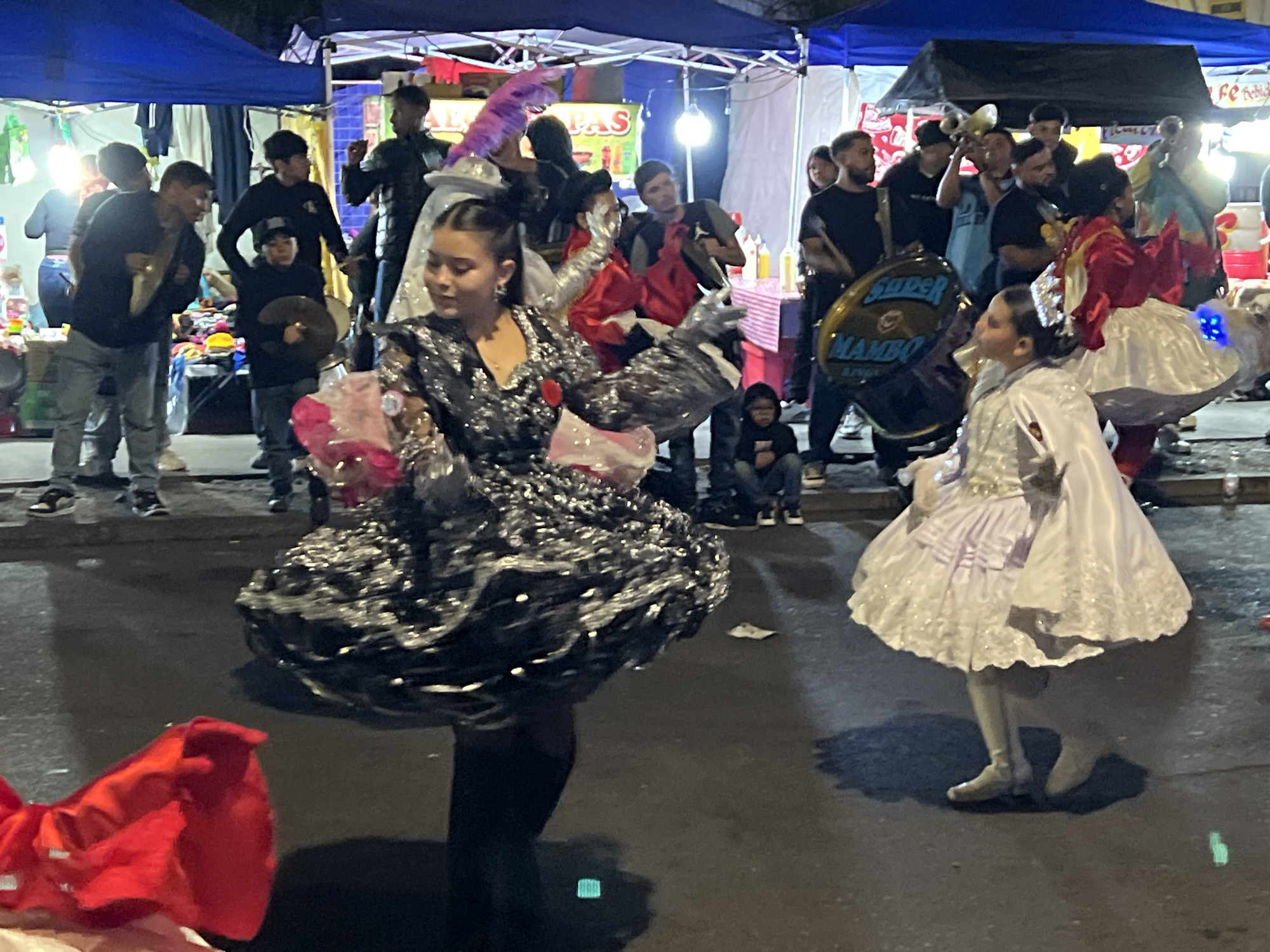In Arica, Chile, at night, a street scene unfolds with a parade or performance. In the foreground, two young girls are dressed in elaborate costumes, dancing. The girl on the left wears a shimmering black dress with a full skirt and silver accents, holding pink decorations. The girl on the right is in a white ruffled dress with sheer layers and appears to be wearing white boots. Behind them, a diverse crowd watches. Several individuals are holding musical instruments, including drums and brass instruments. Some people are standing and observing, while others are seated. Booths with tents and banners are set up along the street, suggesting a market or festival setting. The overall atmosphere is festive and energetic, illuminated by artificial lighting.