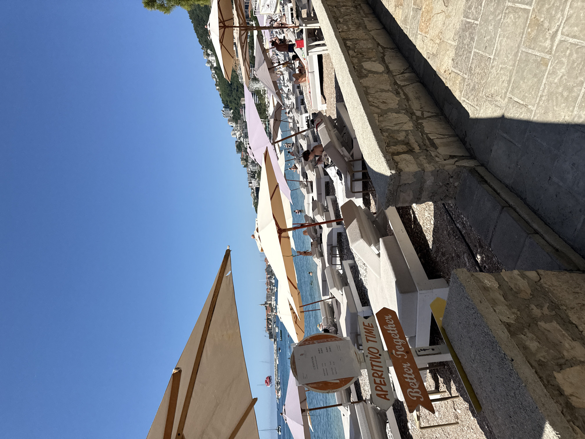 A beach in Budva, Montenegro, is depicted under a clear blue sky. The foreground features a stone wall and a paved walkway. Further back, a section of a beach with numerous white lounge chairs and umbrellas is visible. Two wooden signs are placed on the beach, one reading "APERITIVO TIME" and the other "Better Together." In the background, the Adriatic Sea stretches out, with several boats sailing on it, including a parasailer in the distance. Beyond the sea, the cityscape of Budva rises, characterized by buildings and a church steeple, all nestled against a green, hilly landscape. A few individuals are scattered among the lounge chairs, some relaxing and others engaged in activities. The overall scene conveys a relaxed and sunny atmosphere.