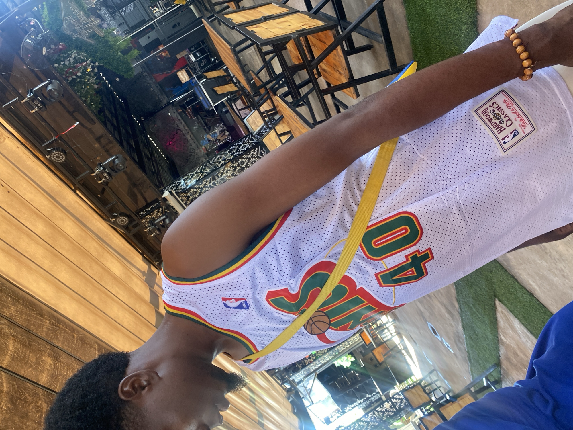 An image taken in what is stated to be Howayi, Nigeria, depicts a dark-skinned male from the chest up, positioned at an angle, within a semi-enclosed commercial venue. The individual wears a white perforated basketball jersey with green, yellow, and red trim around the collar and armholes. The jersey features the NBA logo on the left chest and a graphic with a basketball above the word "HAWKS" (partially visible) and the number "40" in green with a red outline. A "Hardwood Classics Mitchell & Ness" tag is visible near the bottom hem of the jersey. A yellow fabric strap crosses his chest from his right shoulder to his left hip. On his right wrist, he wears a bracelet composed of light brown wooden beads. His hair is short, dark, and curly, and he has a short beard.
The environment appears to be an open-air or sheltered event space, possibly a restaurant or bar. The ceiling is constructed from light brown slatted or corrugated material. Black metal framework supports various fixtures, including LED spotlights and a white oscillating fan. Artificial greenery is suspended from the framework on the right. In the background, a sign partially reads "Road House." A stage area is visible, equipped with musical instruments including a drum set, large black speakers, and a keyboard stand or console. Numerous square tables with wooden tops and black metal frames, along with matching chairs, are arranged across the concrete floor. Sections of artificial green turf are laid out in patterns on the floor. Sunlight illuminates parts of the scene, creating bright spots and glare. A blue garment is partially visible in the bottom left corner of the frame.