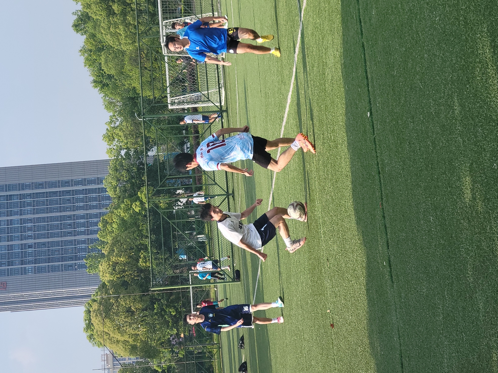 The image shows a group of young men playing soccer on a well-maintained artificial turf field. One player, dressed in a white t-shirt and dark shorts, is kicking the ball. Another player, wearing a light blue jersey with the number 12, is running towards the goal. A third player, wearing a blue jersey, is also on the field. A fourth player, also wearing a blue jersey, is standing near the sidelines. In the background, a soccer goal and a green fence can be seen. Beyond the fence, there are trees and a tall building. The image was taken in the city of Chongqing High-tech Zone, China.