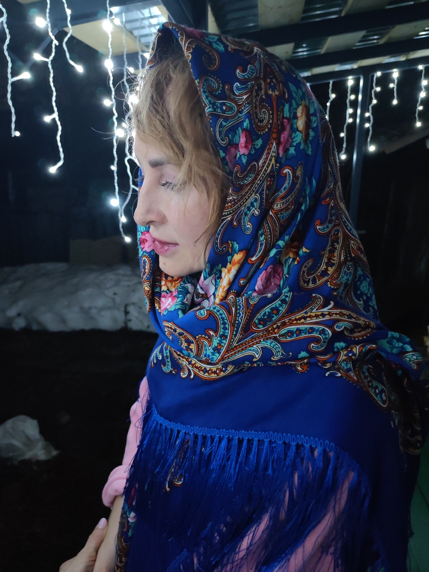 A woman is pictured in profile, facing left, wearing a blue floral patterned headscarf with tassels, and a pink garment underneath. She has blonde hair and closed eyes. The background features a snowy area and string lights. The location is Chaikovsky, Russia.