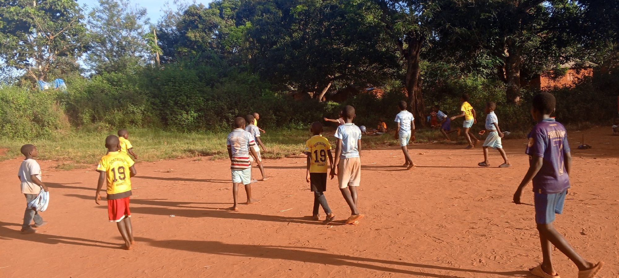 The image depicts a group of children in Ngalula, Democratic Republic of the Congo, on a reddish-brown, dirt-covered ground that appears to be a playing field. The background features trees and green foliage, suggesting a natural or minimally developed environment. At least 11 children are visible, wearing various casual clothes, including t-shirts and shorts; some are wearing soccer jerseys with numbers. Some children are standing while others are running. The light casts long shadows on the ground. In the distance, some structures that might be simple buildings are partially visible.