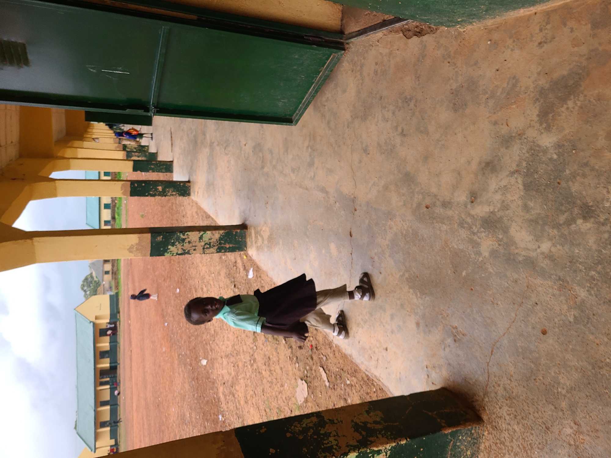 This image captures a young child standing on a concrete walkway, looking directly at the camera, within what appears to be a school compound in Rafin Bauna, Nigeria. The child, likely a girl, wears a light green collared shirt, a dark skirt over light-colored trousers, and open-toed sandals.
The foreground features a covered walkway with yellow pillars and a dark green painted base. A large, dark green panel or door is visible to the right. The concrete floor of the walkway shows some wear and minor cracks. Beyond the walkway, the ground is predominantly reddish-brown dirt with scattered white debris.
In the mid-ground, a long, single-story building with a light-colored facade and a light green corrugated metal roof extends horizontally. Several other smaller figures, presumably children, are visible on the dirt ground near this building, with one person clearly wearing a blue top. Another building is partially visible in the distant background on the left.
The scene is set during the daytime under an overcast sky, providing soft, diffused lighting. The overall environment, with its buildings and children, suggests an educational institution. No visible text is present in the image.