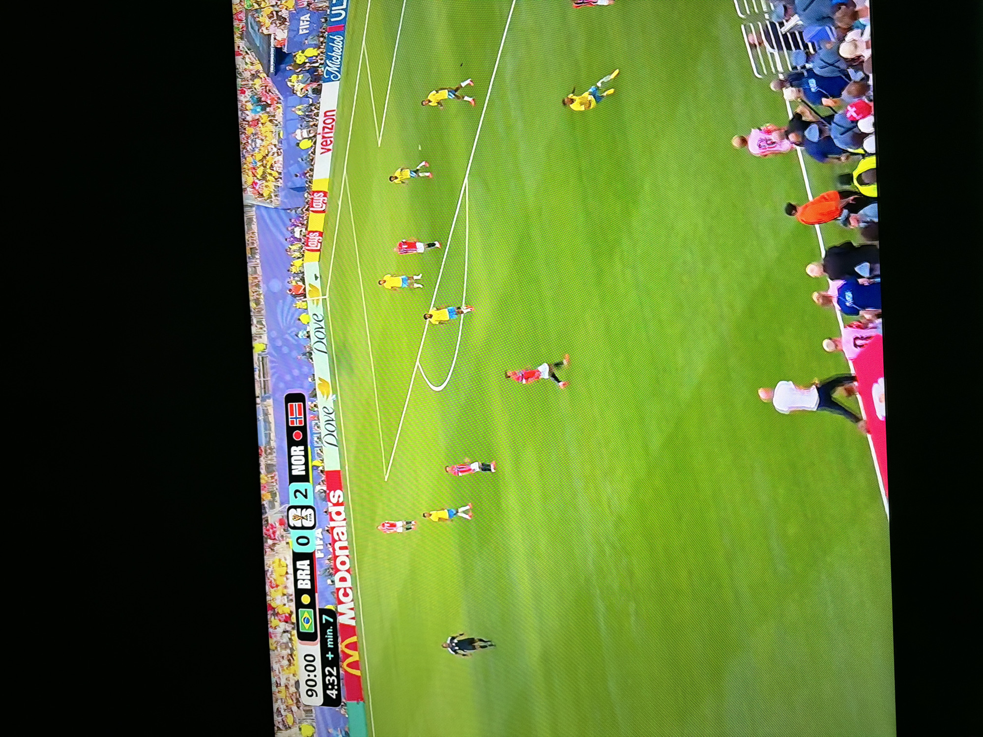 The image shows a soccer match in progress, as indicated by the field markings, players in uniform, and the scoreboard at the top left. The scoreboard reads "90:00" for time and "BRA 0 2 NOR" for the score, with the flags of Brazil and Norway displayed. There are spectators in the stands behind the field, some of whom are wearing yellow and green, suggesting support for Brazil. Advertising boards are visible along the edge of the field, including logos for McDonald's, Dove, and Lay's. The overall setting is a well-lit outdoor sports stadium. The request to include the location in Ognianovo, Bulgaria, cannot be fulfilled as there is no information within the image to support this specific location.
