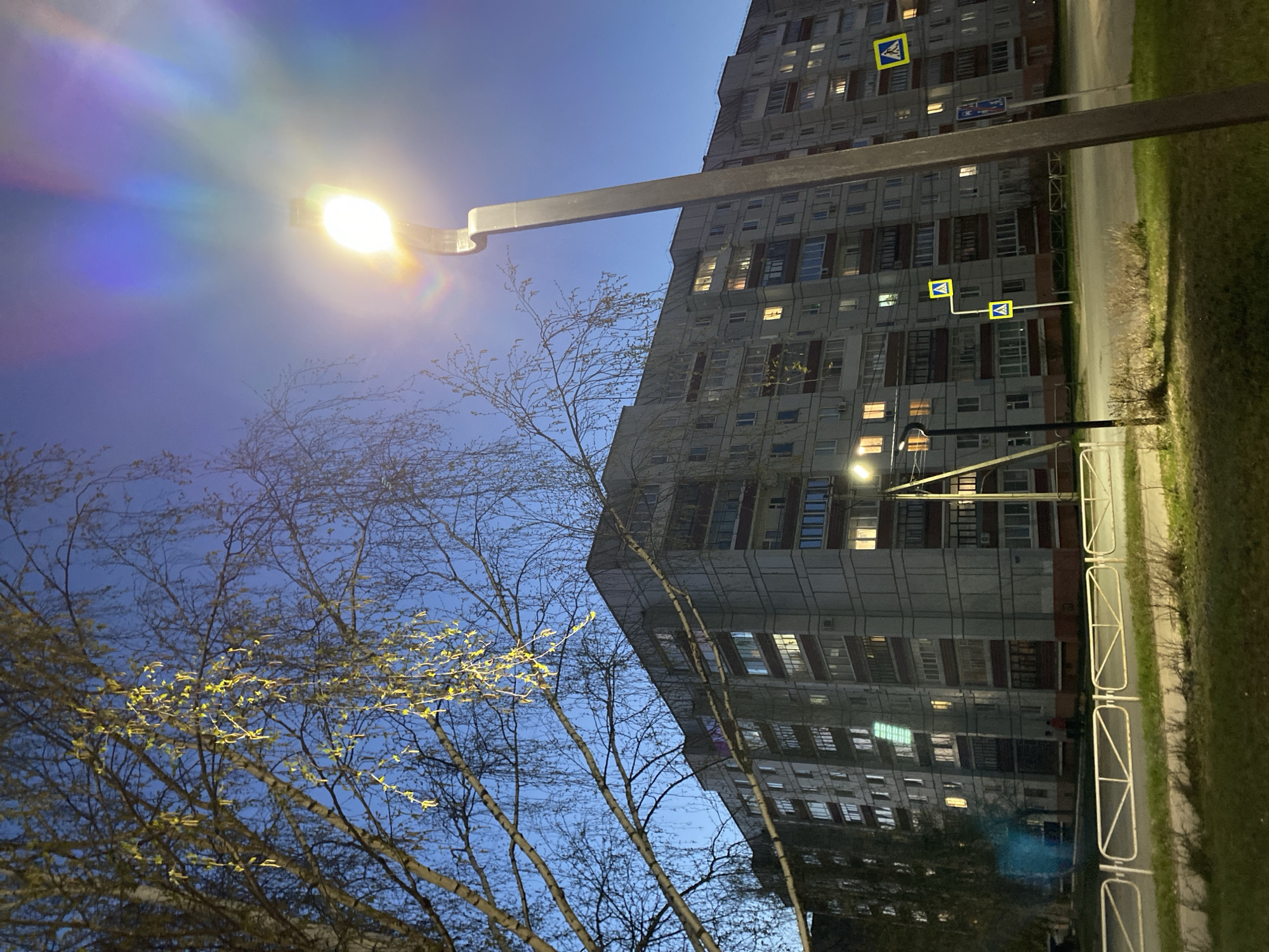 The image depicts a view in the city of Chaikovsky, Russia, at dusk or early evening. A multi-story, gray apartment building with numerous windows, some illuminated, stands prominently. Streetlights with a strong glare illuminate the surroundings and a crosswalk sign. Bare tree branches with early spring buds are visible in the foreground against a blue sky. The ground is covered with green grass. A pedestrian fence is visible, dividing the sidewalk from the grass.
