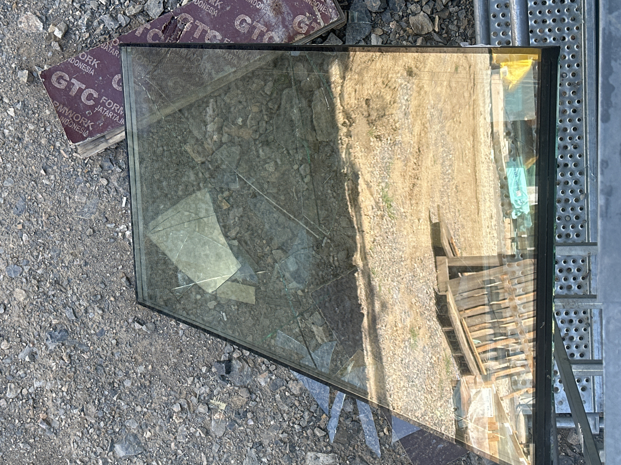 The image shows a broken pane of glass lying on the ground amidst gravel and debris. In the background, a piece of brown cardboard with text including "GTC" and "INDONESIA" is visible. The glass reflects a construction site or an outdoor area with wooden structures and more gravel. The scene appears to be an urban or industrial outdoor setting.