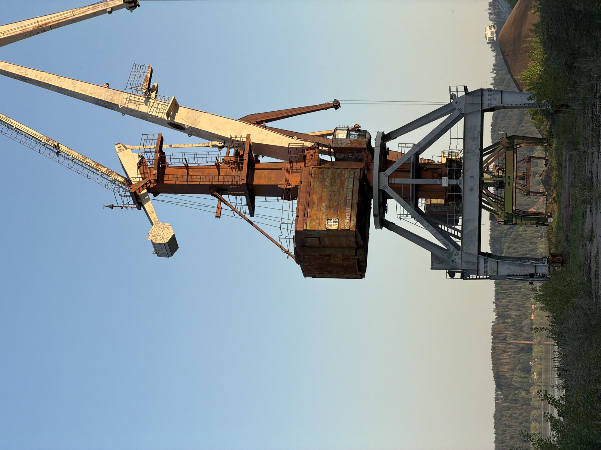 A large, rusted, and seemingly disused harbor crane dominates the foreground, set against a backdrop of a clear sky gradually transitioning to a hazy horizon. The crane’s structural components are painted in light colors that show signs of weathering and rust. The crane's design includes a tall gantry framework with a central hoisting mechanism and a boom arm extending outwards, featuring a cable-suspended weight. The gantry structure is silver/grey with the crane section primarily covered in reddish/brown color. The immediate ground area beside the crane is a mix of foliage and bare earth, leading into a verdant stretch of trees along a body of water. The horizon reveals hints of distant buildings and structures obscured by the atmospheric haze. This is likely located in or around Noviy, Russia, given the crane’s design which is typical of industrial sites in the region.