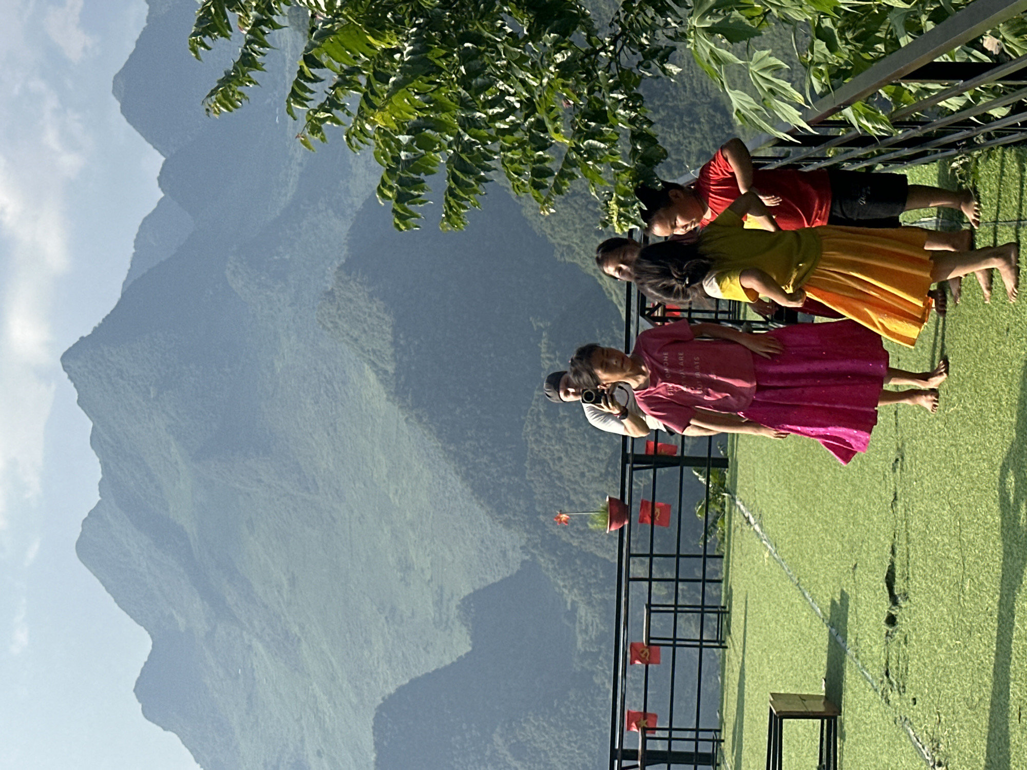 The image depicts a group of four people, seemingly tourists, viewing a mountain range in the Đường Thượng Commune, Vietnam. They are positioned on a balcony or elevated platform with a metal railing, adorned with small red flags. Below, an artificial green lawn extends out. A seated bench and a watering system are visible. One person appears to be taking a photograph of the mountain range. The terrain is mountainous, with lush green vegetation covering the slopes. The sky is partially visible.