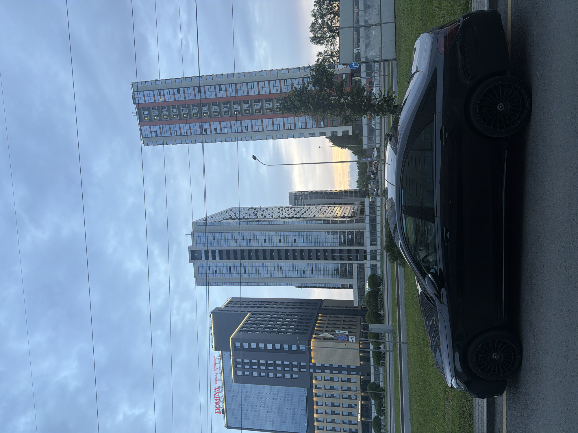 The image shows a black Mercedes-Benz sedan parked on a street in front of several tall, modern buildings. The buildings are made of glass and metal, with a mix of blue and red accents on one of them. The sky is overcast with a few thin clouds. The foreground features the car parked on asphalt with a yellow line marking the edge of the road. Behind the car, there is a grassy median with some bushes and trees. In the distance, more buildings and trees are visible. There are overhead power lines running across the frame.