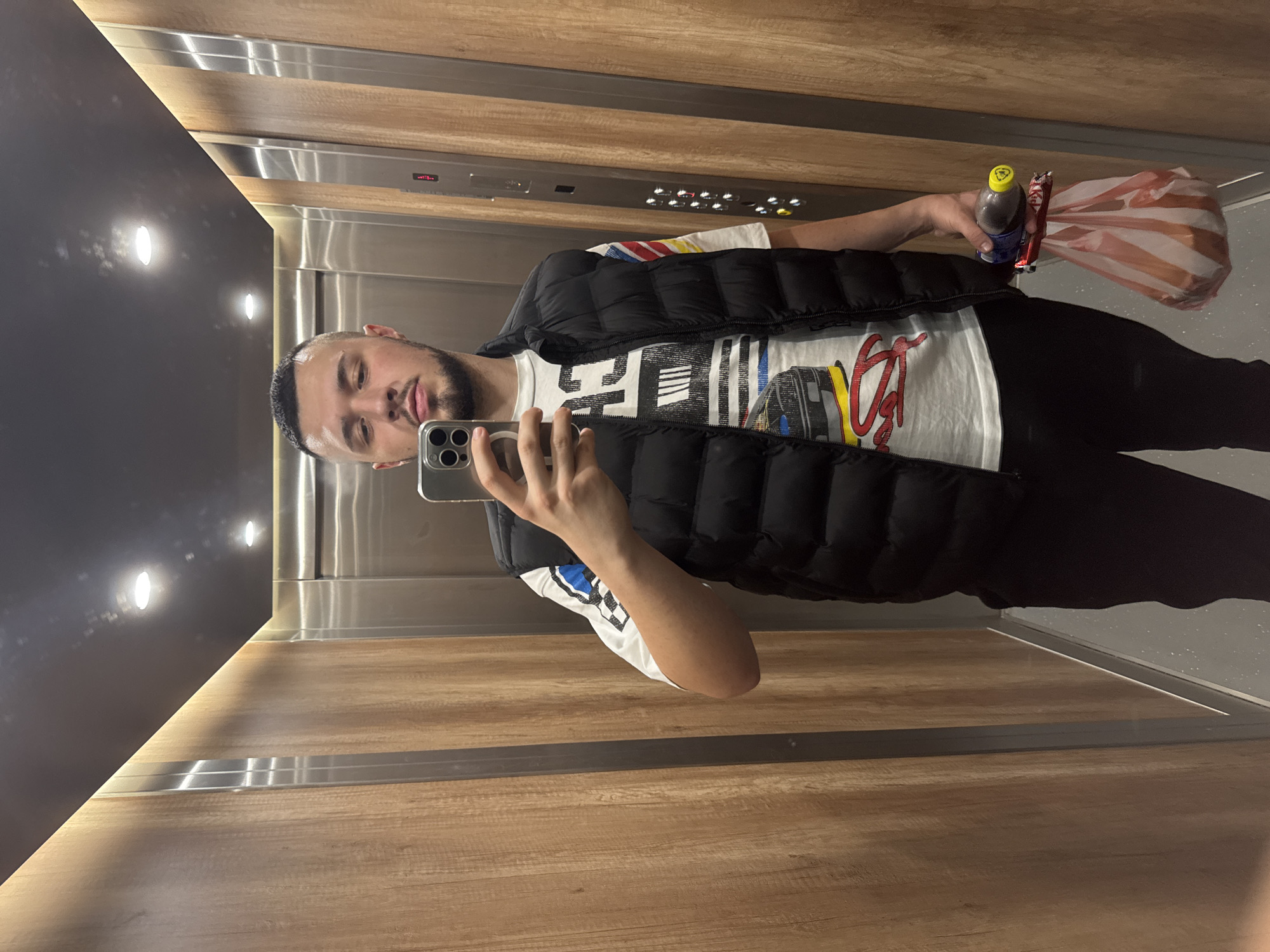 A young adult male is standing in an elevator and taking a selfie with his phone. He is wearing a black puffer vest over a white t-shirt with a graphic print. He has dark hair and a beard. He is holding his phone in his right hand and a plastic bag with items inside, along with a bottle and a candy bar, in his left hand. The elevator walls are paneled with wood and stainless steel. The ceiling has recessed lighting. The elevator floor is a light gray speckled material. The elevator appears to be in a modern building, possibly in Sofia, Bulgaria, based on the context.