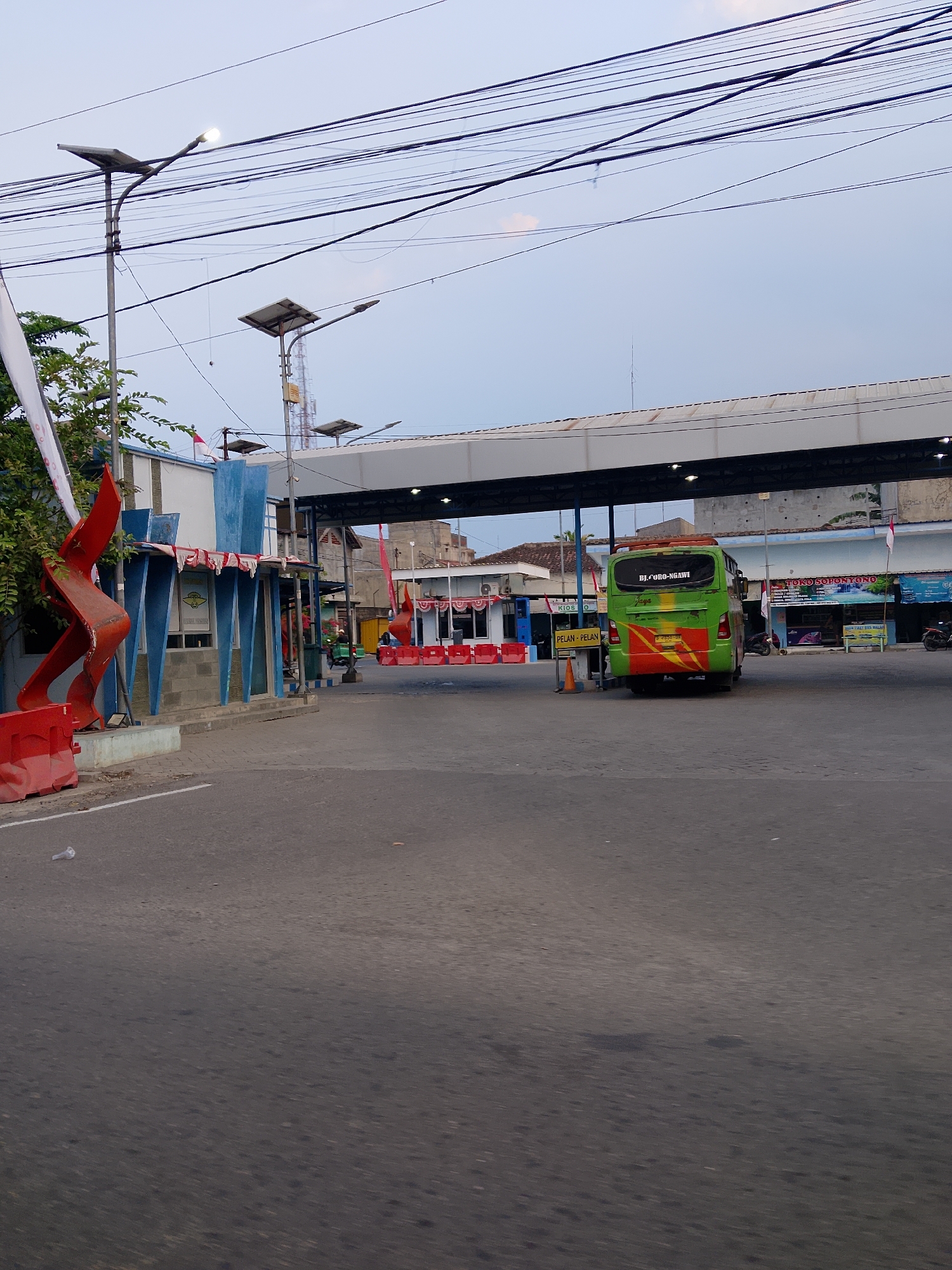The image depicts a street scene in Kuncen, Indonesia, likely near a transportation hub or checkpoint. A green bus with the license plate BJ-080-NGawi is parked under a covered structure. The bus has "Jaga" written on the back in cursive and a red and yellow stripe design. In front of the bus, orange traffic cones are visible, and signs reading "PELAN-PELAN" (slowly) are posted. To the left, a blue and white building with red and white striped awnings is present, with a large red abstract sculpture in front of it. A white banner is also visible on the left. The environment includes overhead power lines and streetlights. The ground is paved, and there is some debris on the road. The sky is overcast.