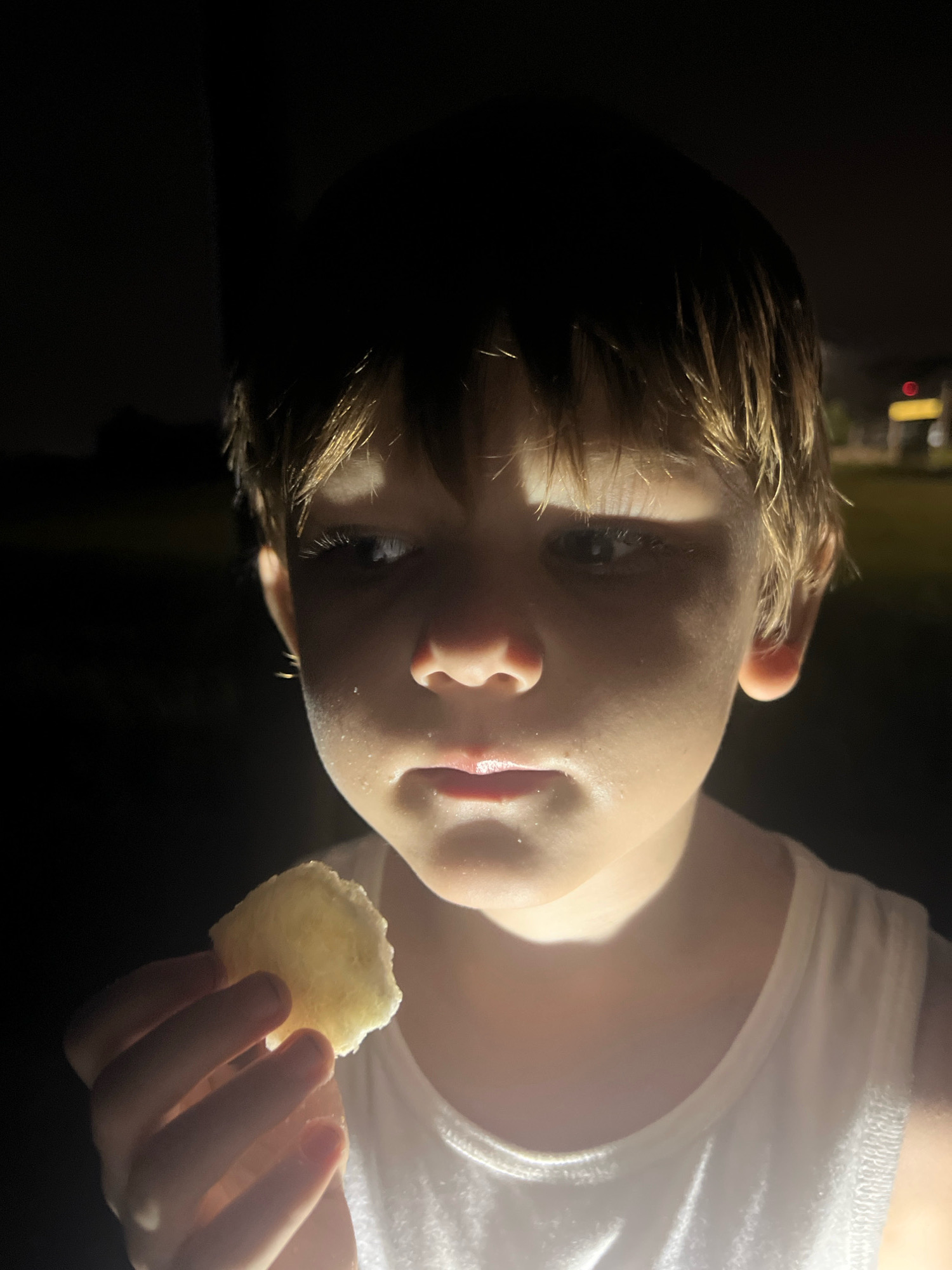 A close-up, low-light portrait shows a young boy, likely around 5-7 years old, holding a piece of food in his left hand near his mouth. The boy is wearing a white sleeveless shirt. He has light brown hair that is somewhat messy. A bright light source, possibly a flashlight or phone, is illuminating his face from the front and slightly below, creating dramatic shadows and highlighting the sweat on his forehead and cheeks. His gaze is directed off to his left, with a slightly pensive or contemplative expression. The background is dark and indistinct, with some faint lights visible in the distance suggesting an outdoor setting at night. There are no discernible location cues for Hoi An, Vietnam, or any visible text.
