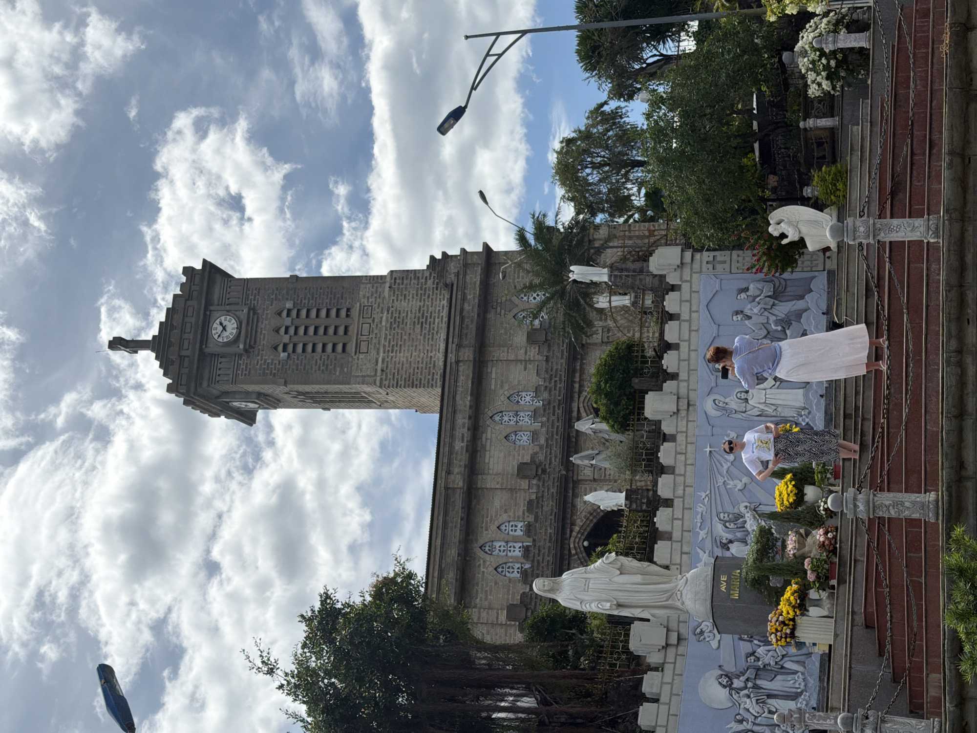 This is a full-shot photograph of the Nha Trang Cathedral in Nha Trang, Vietnam. The cathedral is made of stone and features a tall clock tower. In front of the cathedral, there are several statues, including a large statue of the Virgin Mary. Two women are standing on the steps in front of the cathedral, one of whom is taking a photo of the other. The sky is cloudy.