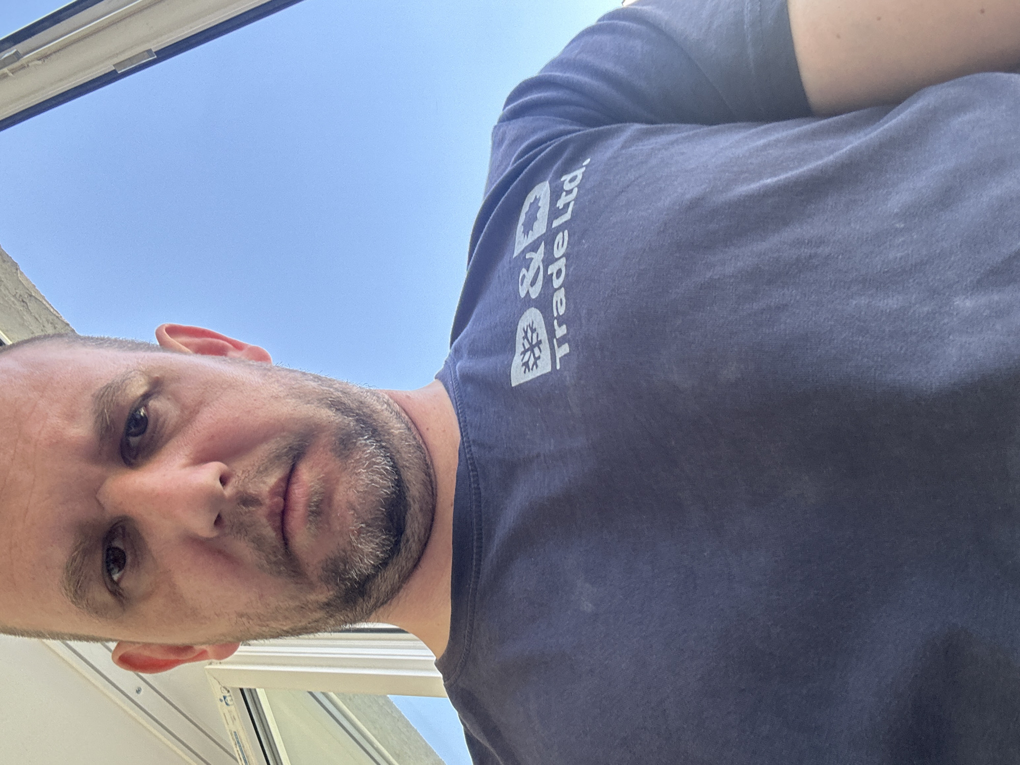 The media file features a man captured from a low-angle perspective, looking downwards directly into the camera. He has fair skin, short dark hair, and a short beard or stubble covering his chin and jawline. His expression is neutral.
He is wearing a dark blue, short-sleeved t-shirt. On the right sleeve, the white text "D & D Trade Ltd." is clearly visible, positioned below two symbols. One symbol resembles a snowflake or an air conditioning unit, while the other is less distinct but similar in style, suggesting a company involved in climate control, heating, ventilation, or refrigeration.
The background is a bright, clear blue sky, indicating daytime and good weather. Parts of a white window or skylight frame are visible in the upper left and lower right sections of the image, suggesting the man is positioned above the camera, possibly through an opening or standing under a skylight. The unusual angle gives the impression of looking up at the subject. The photograph was taken in Sofia, Bulgaria.