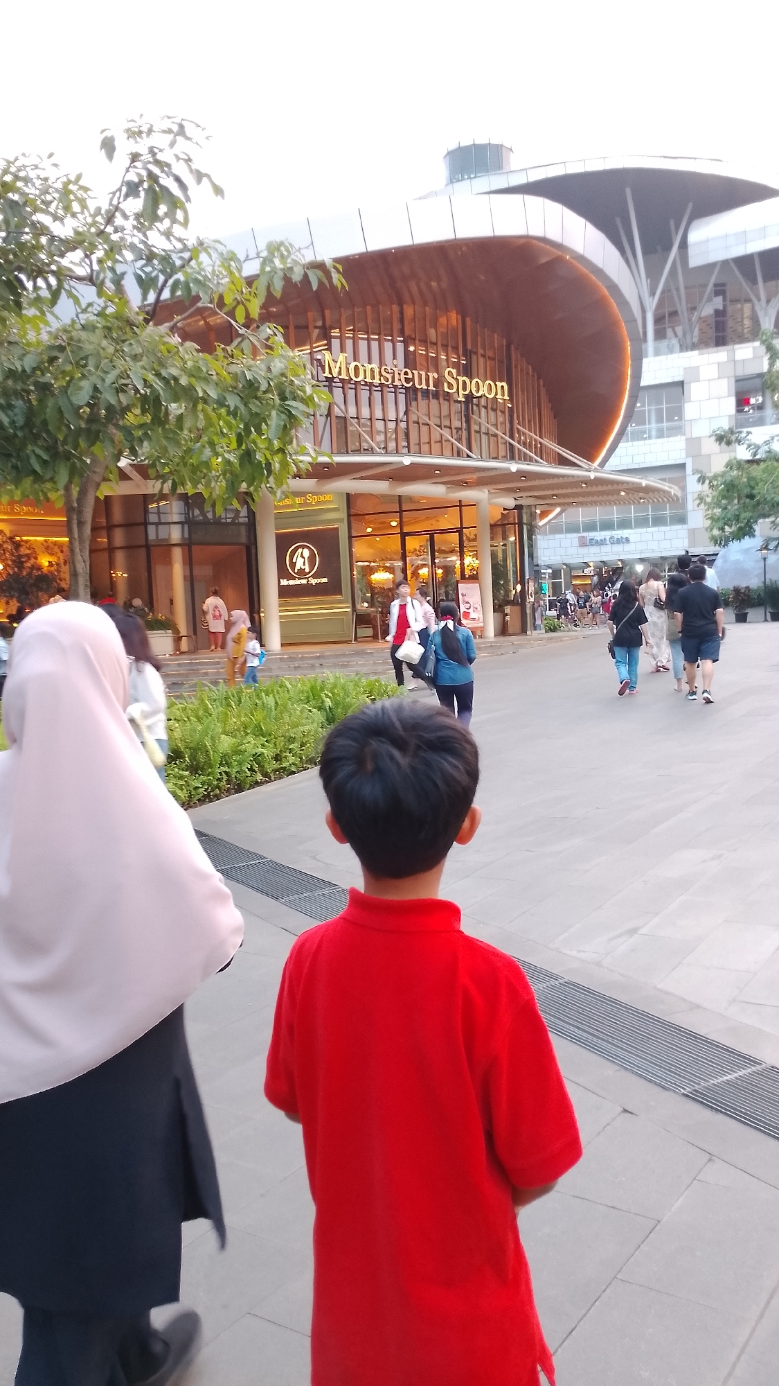 The image depicts the exterior of the Monsieur Spoon restaurant, a popular French-themed eatery located in South Tangerang, Indonesia. The restaurant's facade is characterized by its contemporary architecture, featuring a curved wooden roof structure with glass and wood accents. The prominent "Monsieur Spoon" signage is visible in gold lettering against a warm, illuminated background.
In the foreground, the backs of two individuals are visible. Closest to the camera is a young boy wearing a bright red shirt. To his left and slightly further away is a woman wearing a light pink hijab and a dark outfit. They are walking away from the restaurant, towards the viewer.
In the mid-ground, several other people are seen walking on the paved walkway in front of the restaurant. Some are entering or exiting the establishment, while others are passing by. The area is landscaped with lush green bushes and trees, contributing to a pleasant urban environment. The overall scene suggests a busy public space, likely a commercial or entertainment district, with people enjoying an evening outing. The lighting indicates it is likely late afternoon or early evening, with artificial lights within the restaurant and ambient natural light creating a warm glow.