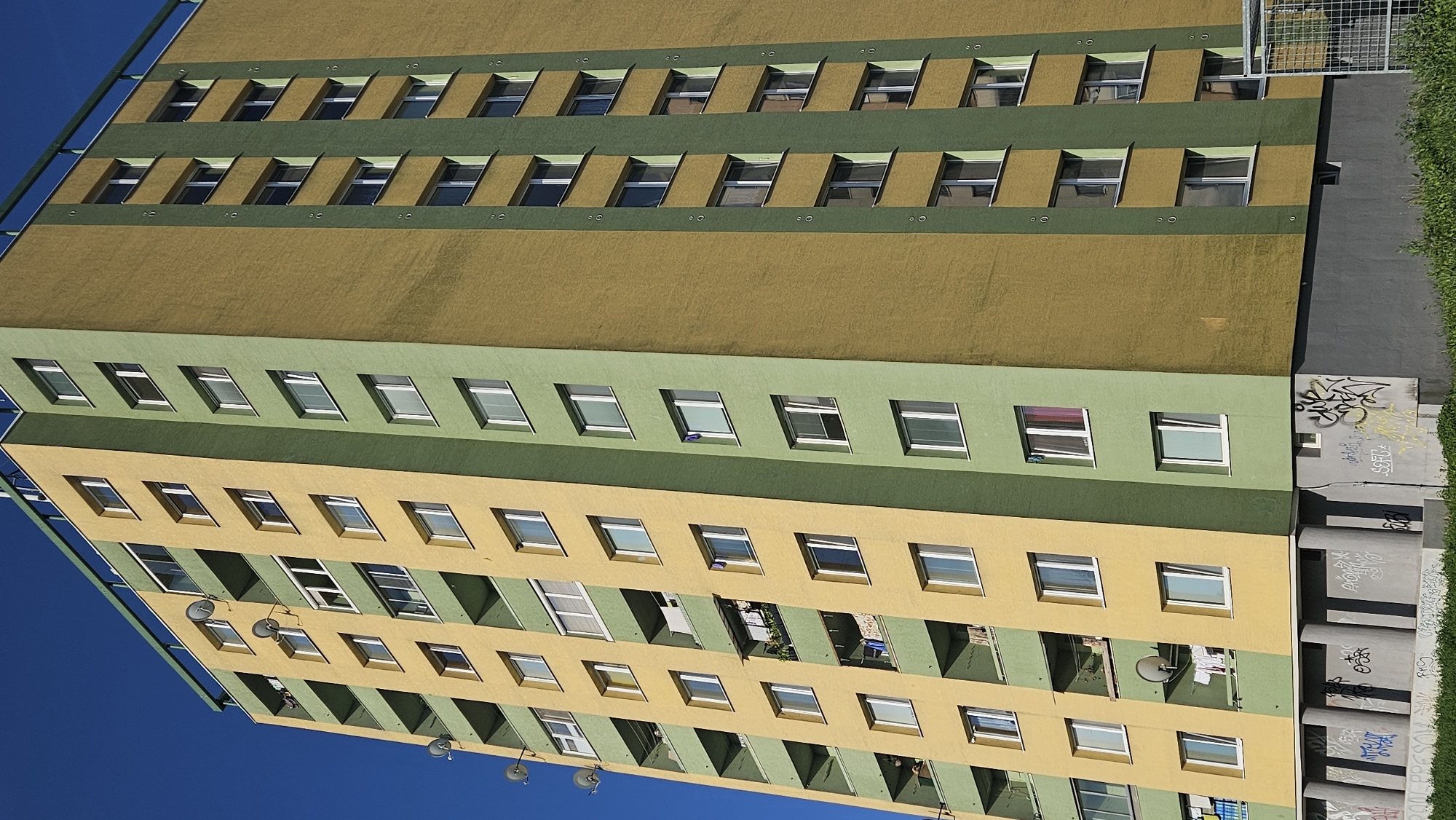 The image shows a tall, multi-story apartment building in the city of Prešov, Slovakia, under a clear blue sky. The building's facade is painted in alternating vertical sections of bright yellow and muted green. It features numerous windows and balconies. Several satellite dishes are visible on some of the balconies. The ground floor of the building appears to be a darker grey color and is adorned with graffiti. In front of the building, there is a patch of green grass and vegetation. To the right of the building, a metal cage structure is partially visible. No people are clearly depicted in the image.