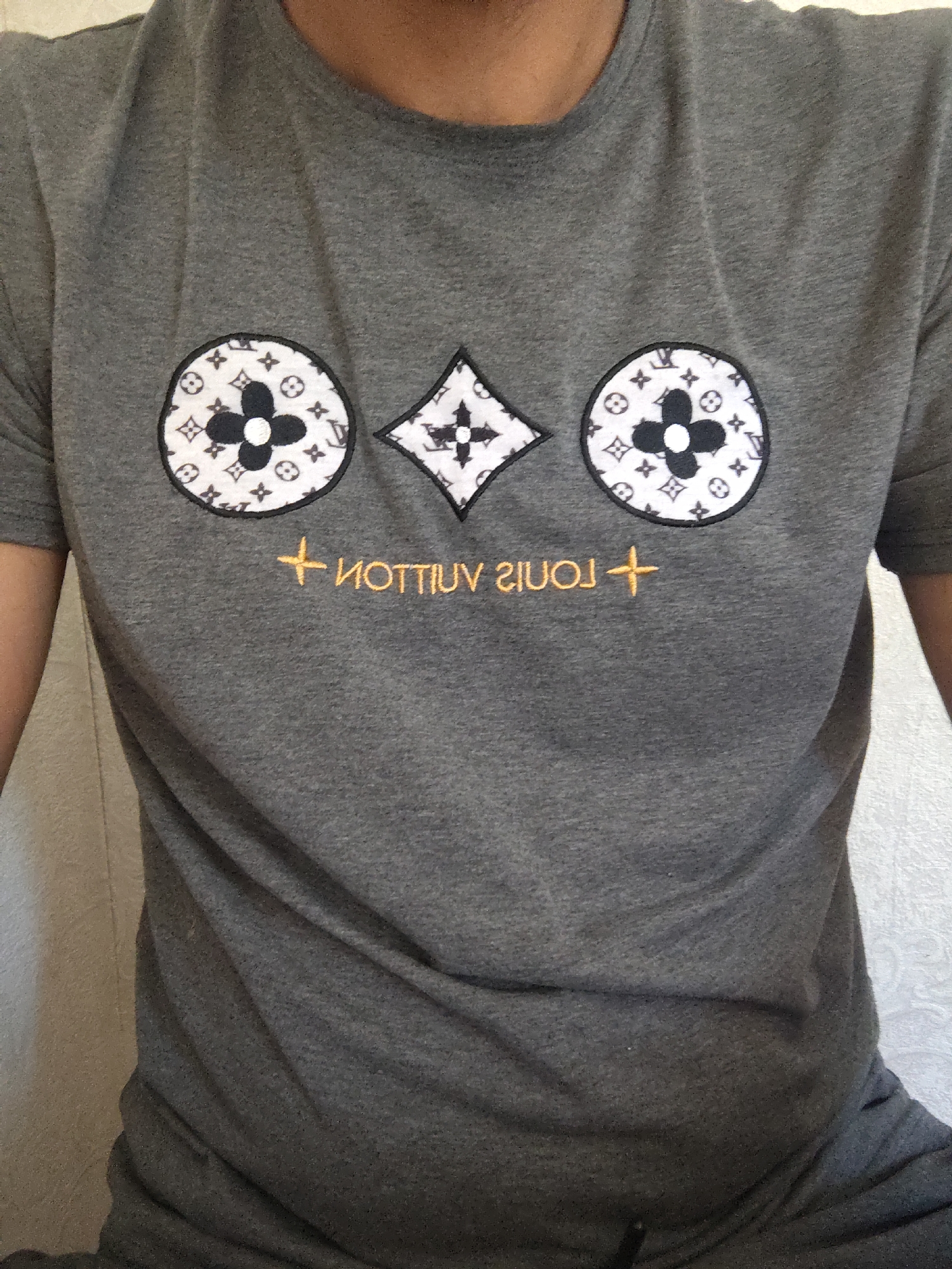 The image shows a person wearing a grey t-shirt with a graphic design on it. The design features three emblems arranged horizontally: a circular emblem on the left with a black four-petal flower inside a white circle patterned with smaller icons, a diamond-shaped emblem in the center with a black and white checkered pattern and a small black cross in the middle, and another circular emblem on the right similar to the one on the left. Below the emblems, text appears to be embroidered in an orange-yellow color, reading "NOTTIV LUOJ" which is "LOUIS VUITTON" spelled backward, with a cross symbol to the left of the text. The t-shirt is made of a soft, textured fabric. The background appears to be a lightly textured wall, possibly in a residential setting. The lighting suggests an indoor environment. No specific location in the city of Novomikhaylovsky, Russia can be determined from the visual information provided.