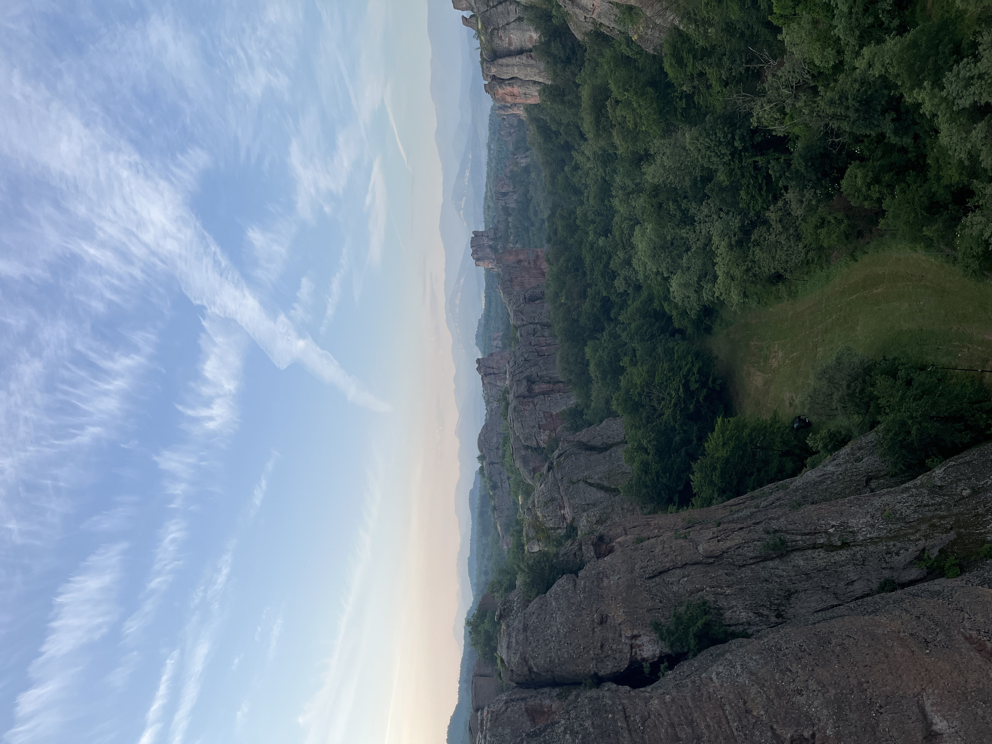 A high-angle, vertical landscape photograph captures the majestic formations of the Belogradchik Rocks in Belogradchik, Bulgaria, under a clear, pale blue sky with a gradient transition to lighter tones near the horizon, characteristic of early morning or late evening light. The sky features delicate, elongated cirrus clouds spread across the upper portion and a prominent white aircraft contrail diagonally traversing from the mid-left towards the upper-right. The midground and background are dominated by massive, reddish-brown sandstone rock formations of varying heights and shapes, interspersed with and partially covered by a dense, verdant forest. Multiple layers of hazy, blue-tinted mountains are visible in the far distance, receding into the atmospheric perspective. The immediate foreground is framed by imposing, rugged rock faces on the left and right. At the bottom-center of the image, amidst the trees, a narrow, grassy clearing contains a dark-colored vehicle. No human figures or animals are present in the scene.