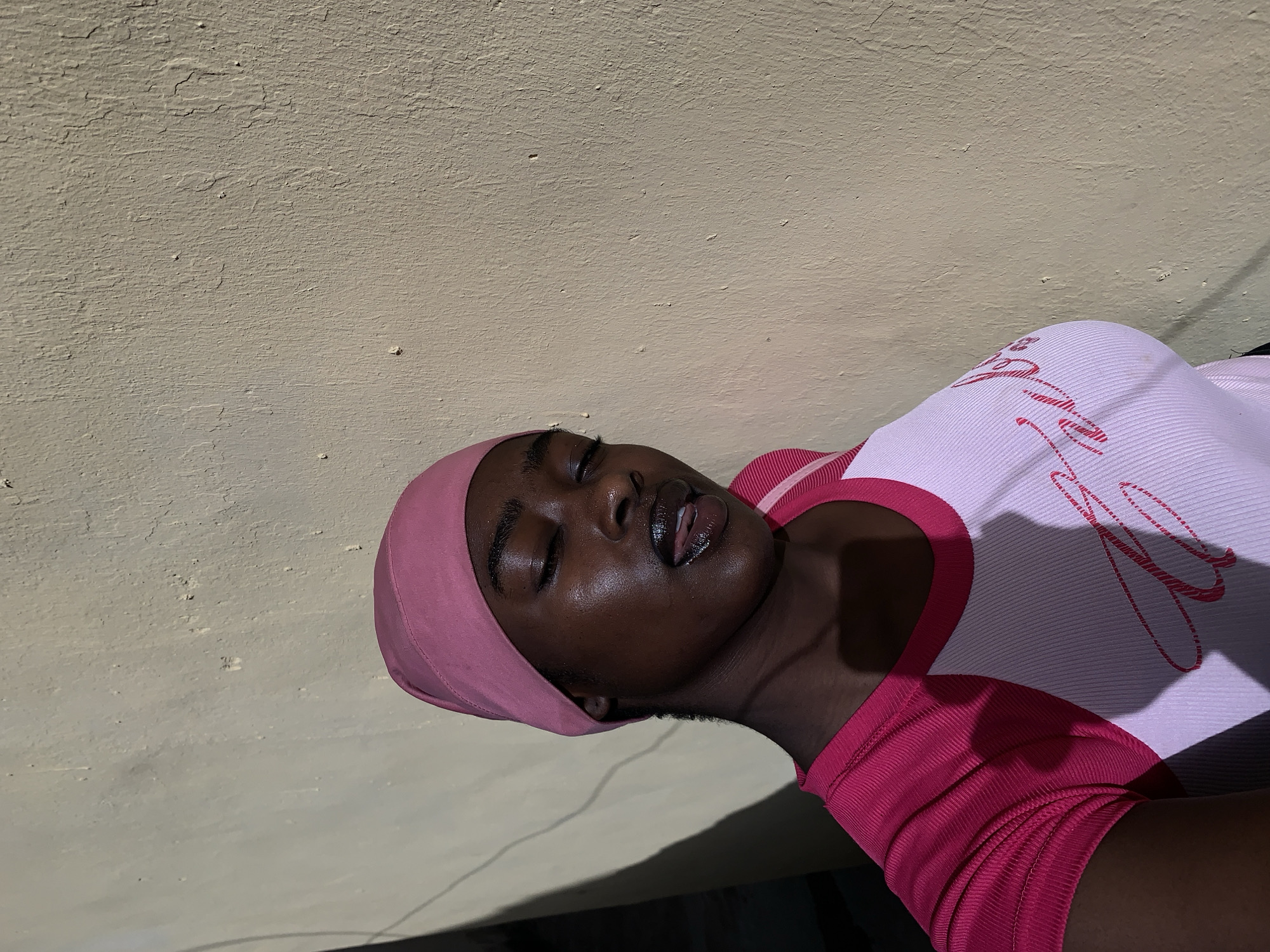 A dark-skinned woman wearing a pink headscarf and a pink and white top is lying down against a beige wall. The photo is taken from a high angle, with the woman's head near the left side of the frame. Her eyes are closed, and her lips are slightly parted. The top has some writing on it. A shadow falls across her chest and the wall. The photo appears to be taken outdoors in Rafin Par, Nigeria, where the lighting is bright.