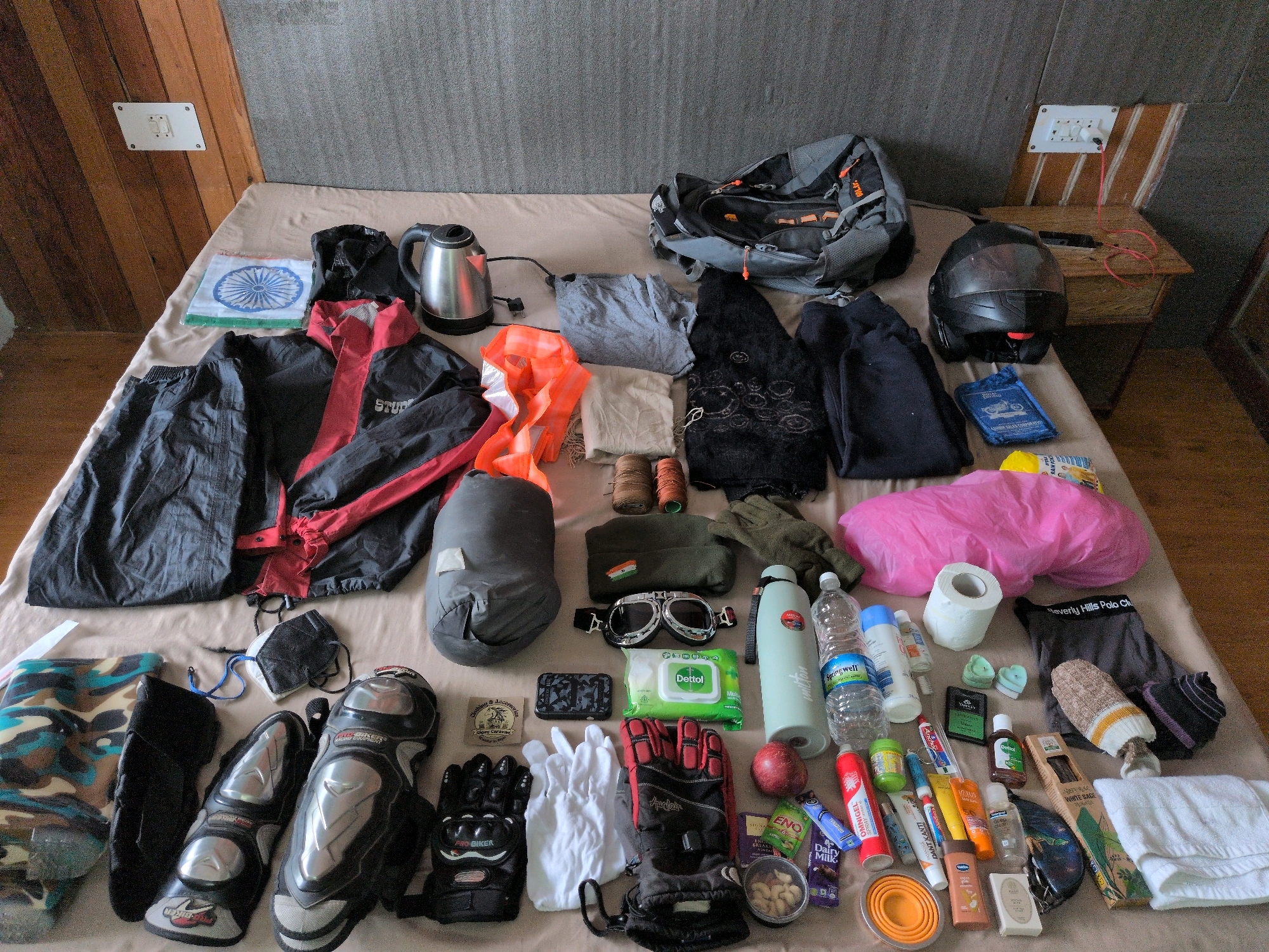 The image displays a collection of travel gear laid out on a bed in Vashisht, India. The scene is shot from a high angle, capturing a wide assortment of items organized on a neutral-colored bedspread. Dominating the top right is a large, dark gray backpack with orange accents. To its left sits a black motorcycle helmet, resting on a wooden bedside table with visible electrical outlets and wiring.
On the bed itself, an array of clothing and accessories are spread out. A black and red motorcycle jacket is prominently placed on the left, with a gray sleeping bag or similar cylindrical item next to it. Other clothing items include dark trousers, black leggings, and a black embroidered garment. Safety gear is also evident, with black and silver motorcycle knee guards and gloves laid out.
Various personal care and travel essentials are present. A silver electric kettle, a roll of toilet paper, and several bottles of water and other liquids are scattered across the bed. Small items like packets of wet wipes, toothpaste, mouthwash, and soap are also visible. A collection of snacks, including chocolate bars and a small container of nuts, are arranged in the lower right quadrant.
Decorative elements include a small white cloth with a blue Indian flag design. The overall impression is one of preparation for a journey, likely involving motorcycle travel, given the presence of the helmet and riding gear. The room appears to be a simple accommodation, possibly a guesthouse or hotel room, with wooden paneling on the wall behind the bed.