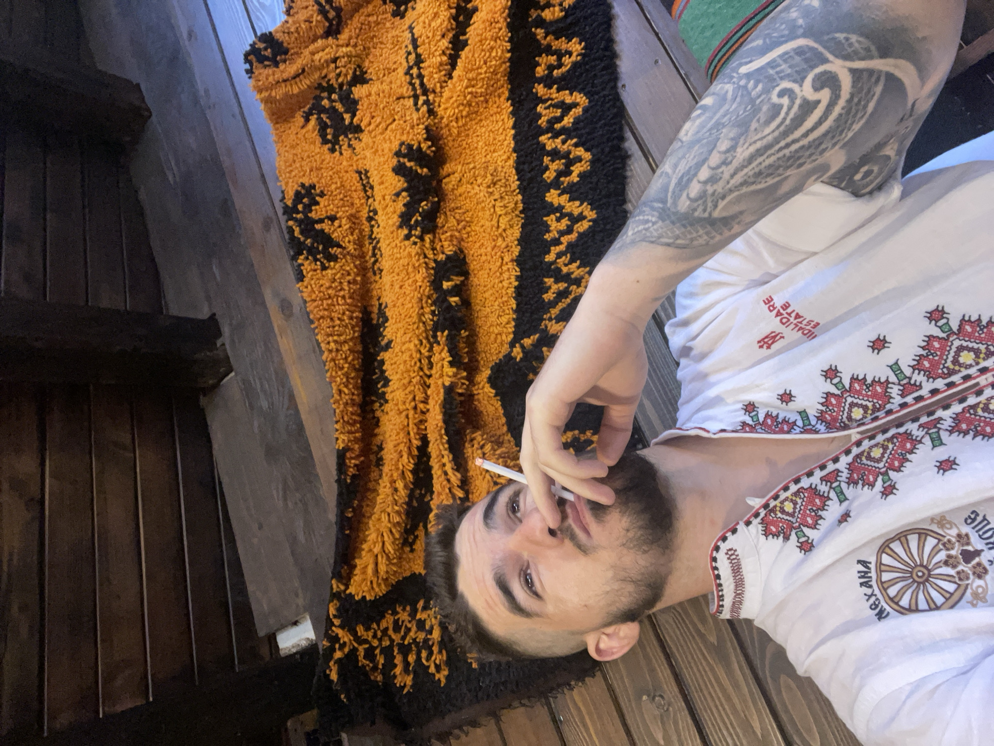A man with a beard and a fade haircut is lying down on a wooden floor with a rug. He is wearing a white shirt with colorful embroidered designs. He is holding a cigarette in his right hand and looking up. The rug is orange and black and has a geometric pattern and some tree-like designs. The background consists of wooden planks.