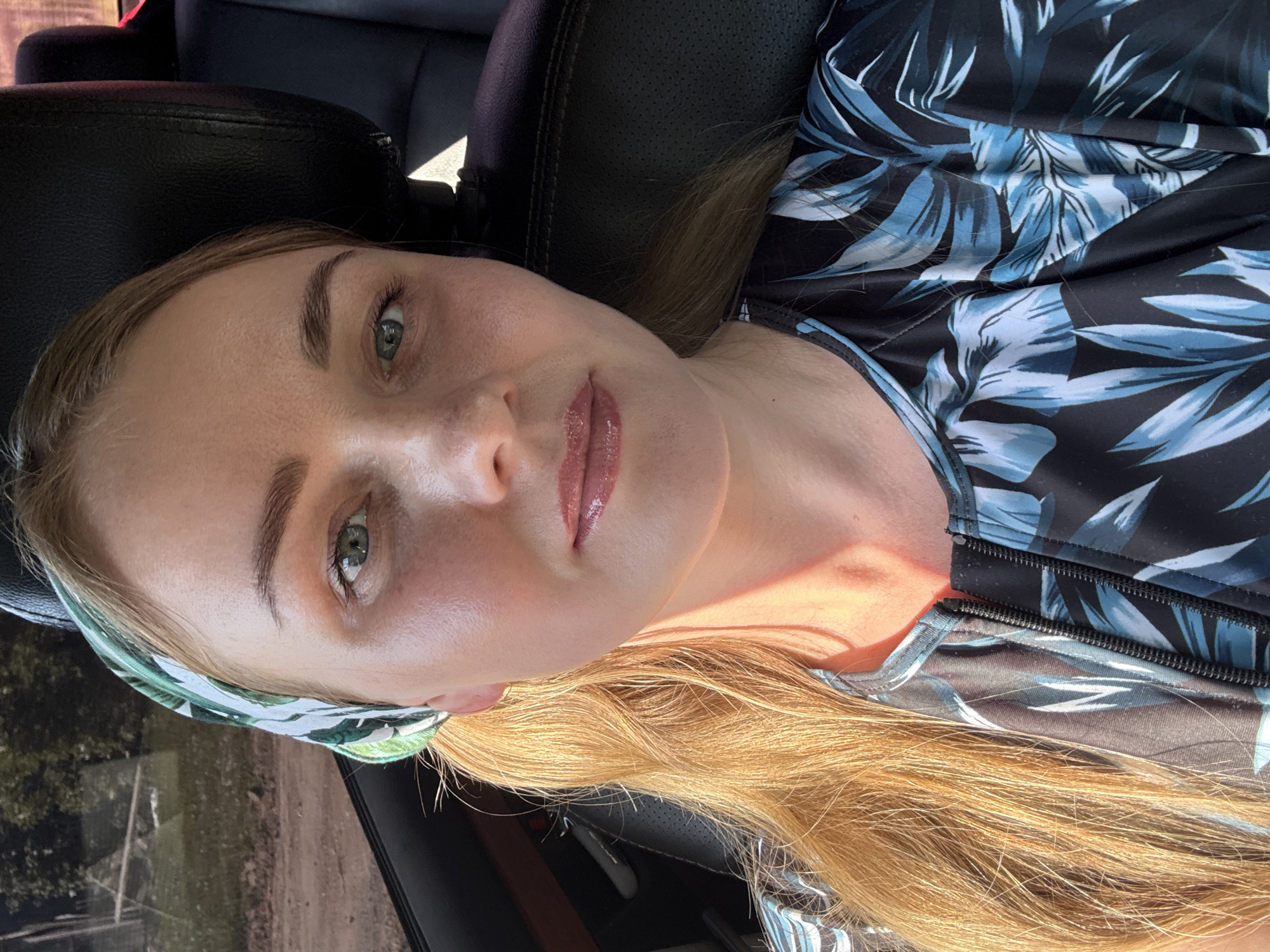A fair-skinned woman with blond hair and light green eyes, wearing a black, blue and white floral patterned top and a green and white patterned headband, is sitting in a car. The car's seat is black leather. The location is unknown, however, based on the prompt it should be Chaikovsky, Russia. The woman is looking at the camera.